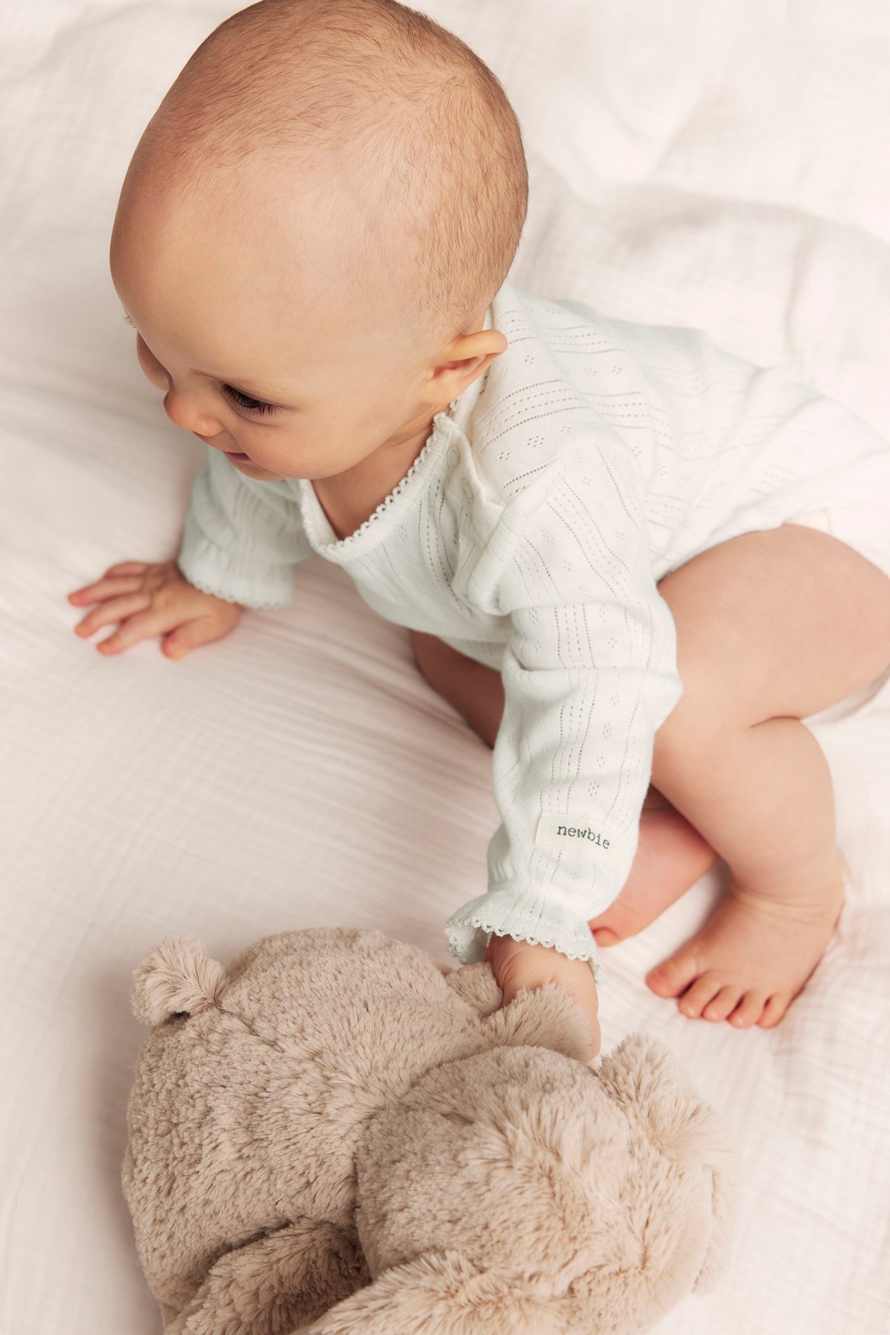 Top-down view of a baby in an offwhite pointelle ribbed bodysuit with ruffled cuffs, touching a beige teddy bear.