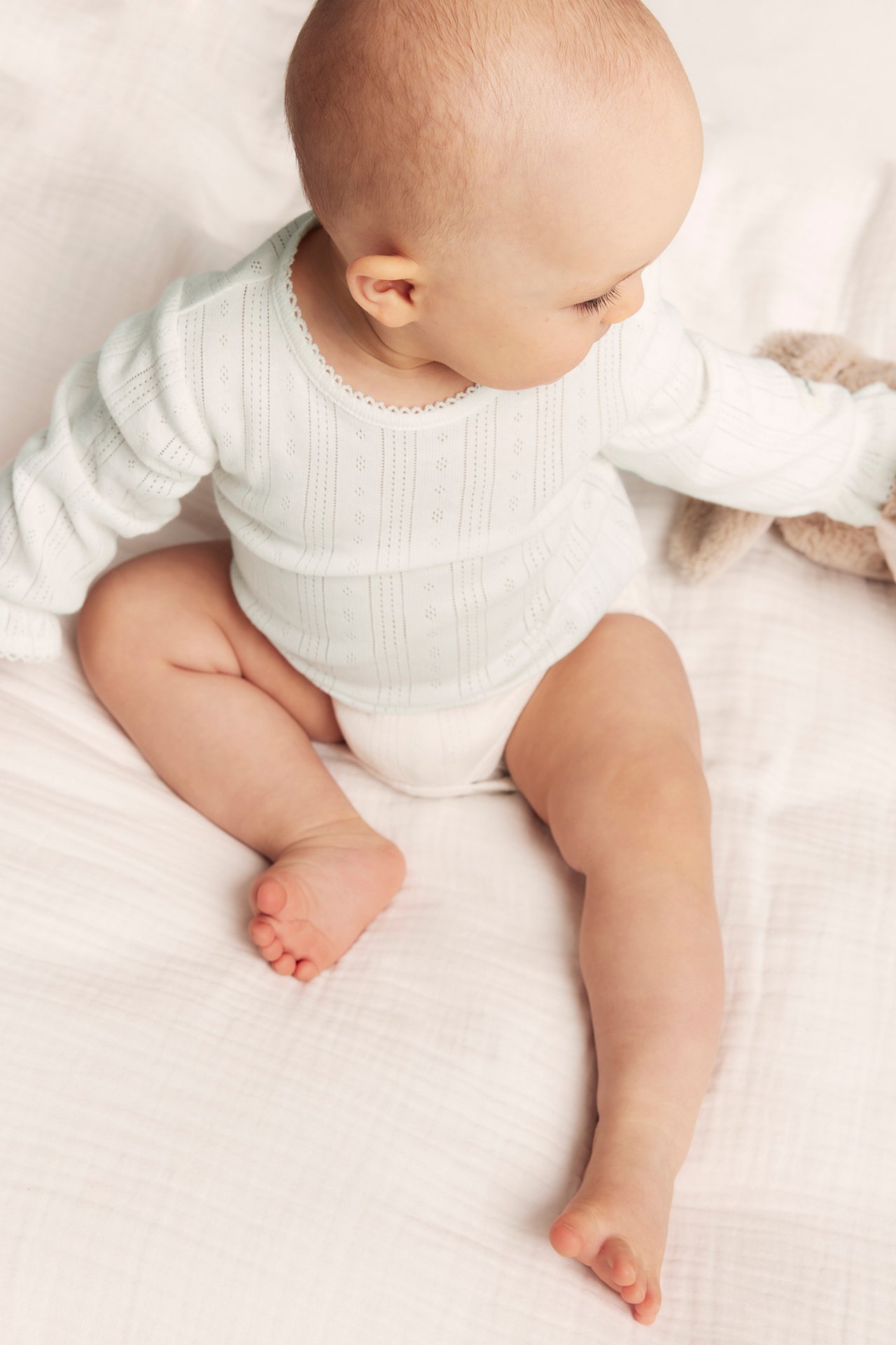 High-angle view of a baby in an off-white pointelle ribbed bodysuit with picot trim, holding a plush toy.