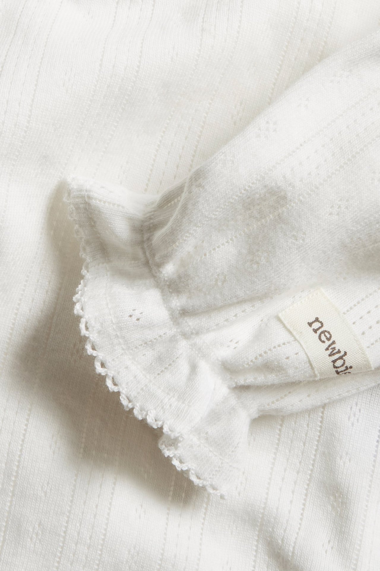 Nahaufnahme eines weißen gerippten Babykleidungsstücks mit Lochstickerei, Rüschenbesatz und Newbi-Etikett.