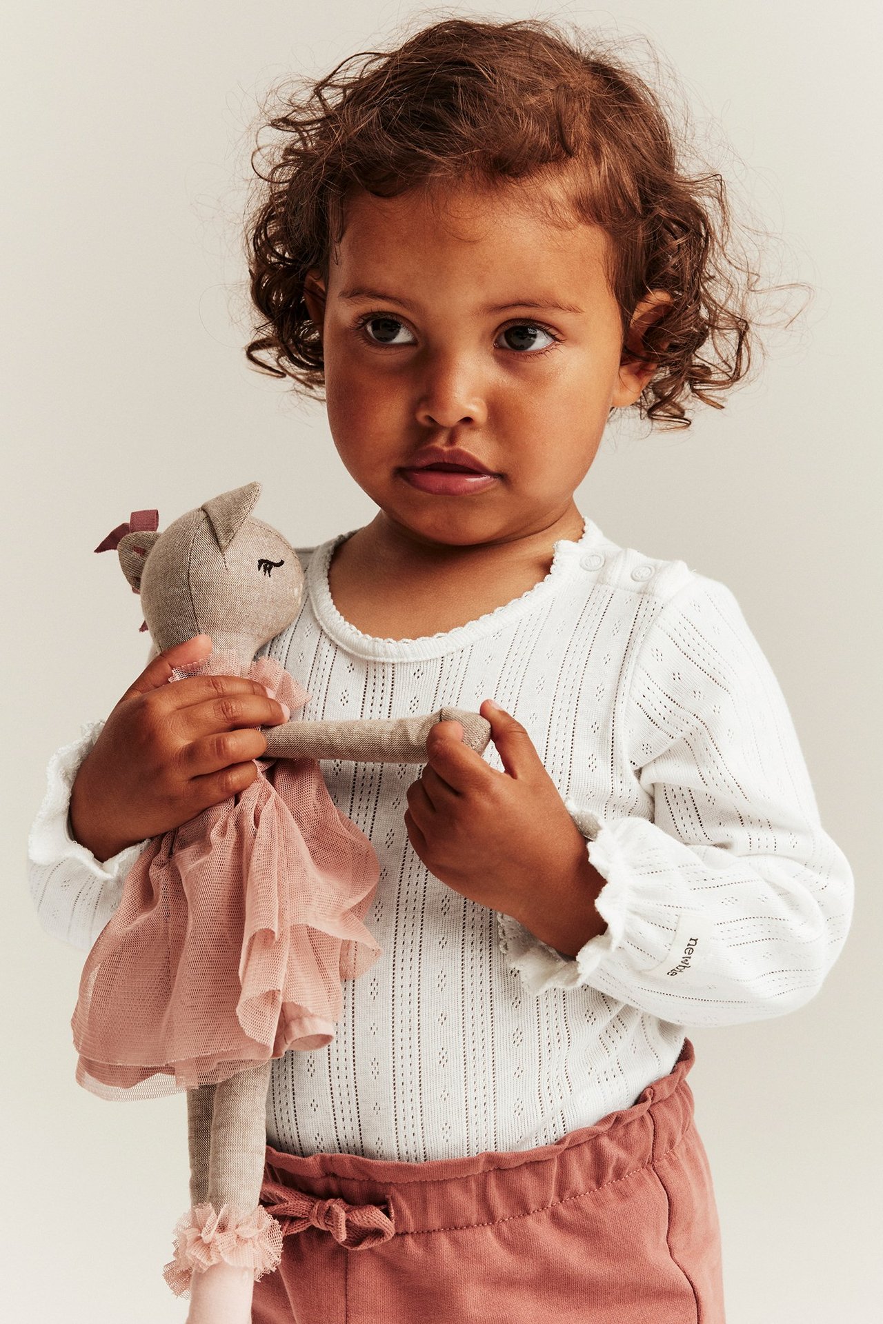 Vorderansicht: Kind in cremeweißem Pointelle-Ripp-Langarmbody mit Rüschenärmeln, rosa Hose, hält eine Puppe.
