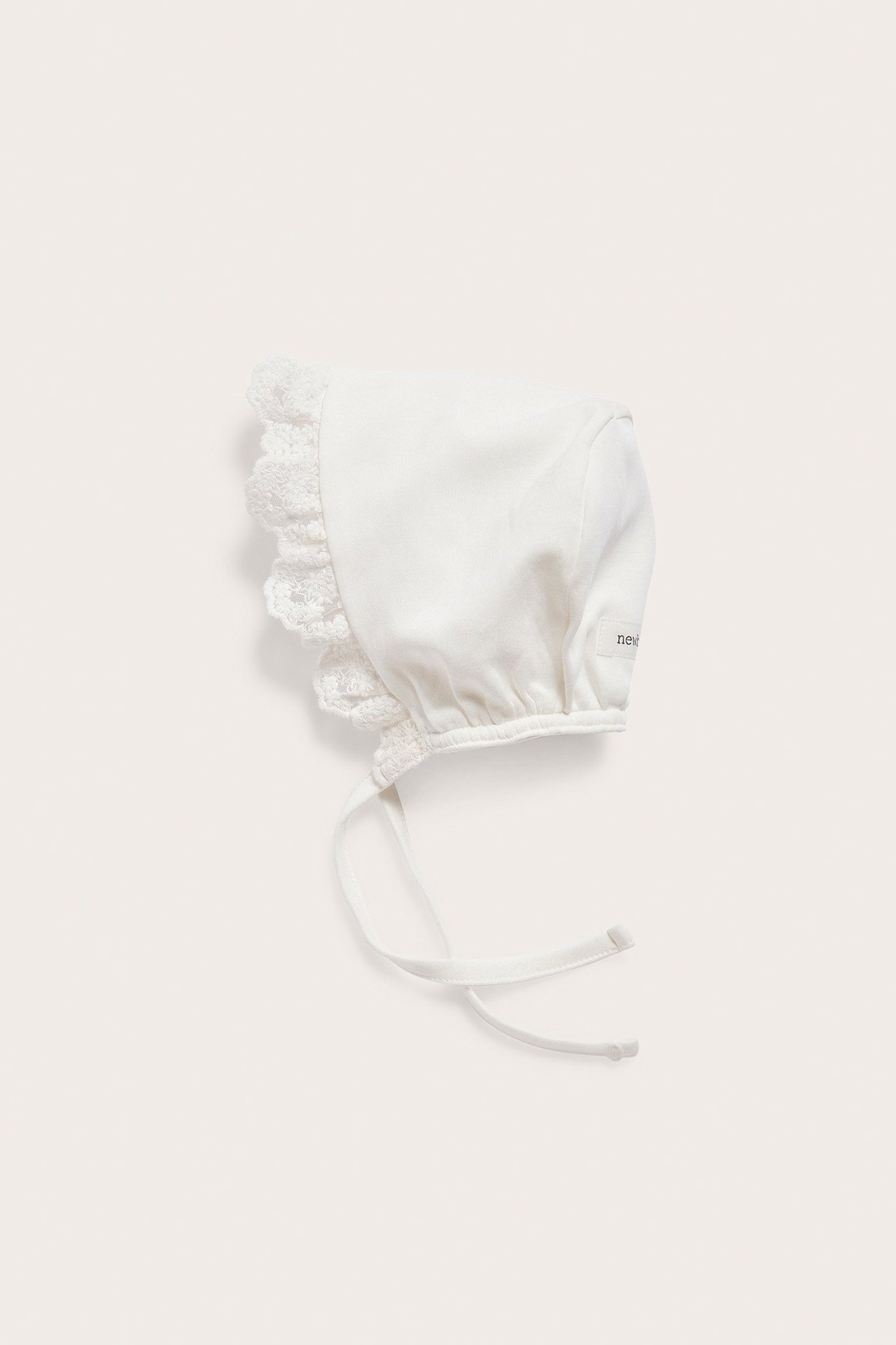 Side view of an offwhite baby bonnet in soft jersey with a lace frill and tie bands.