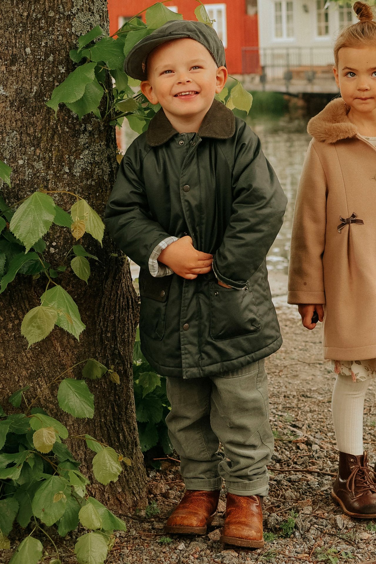 Vorderansicht: Kind in grüner Wachsjacke, Mütze, Hose, braunen Stiefeln. Mädchen trägt beigen Mantel.