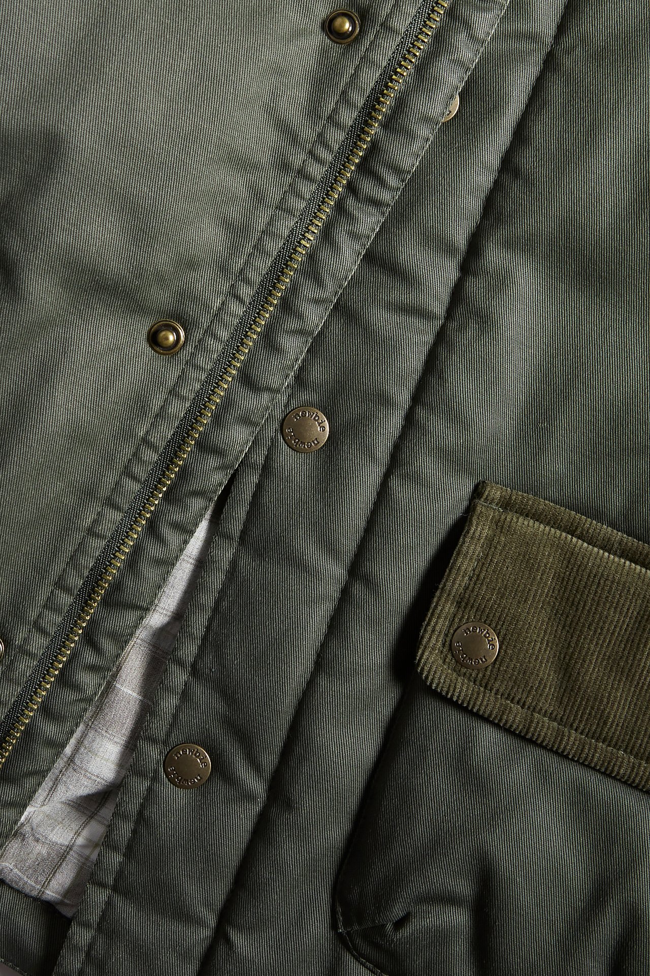 Close-up of a green waxed cotton jacket with zipper, snap buttons, corduroy flap, and patterned lining.