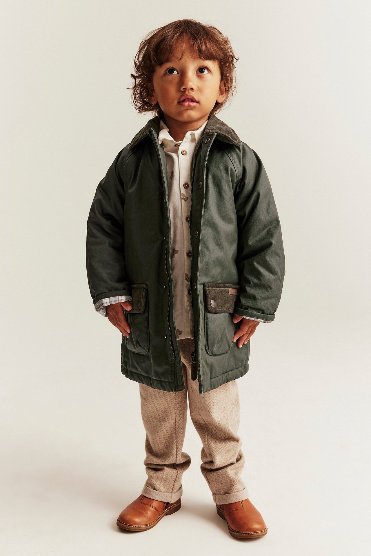 Front view: Kid in green waxed padded jacket with corduroy collar, patterned shirt, beige trousers, brown shoes.