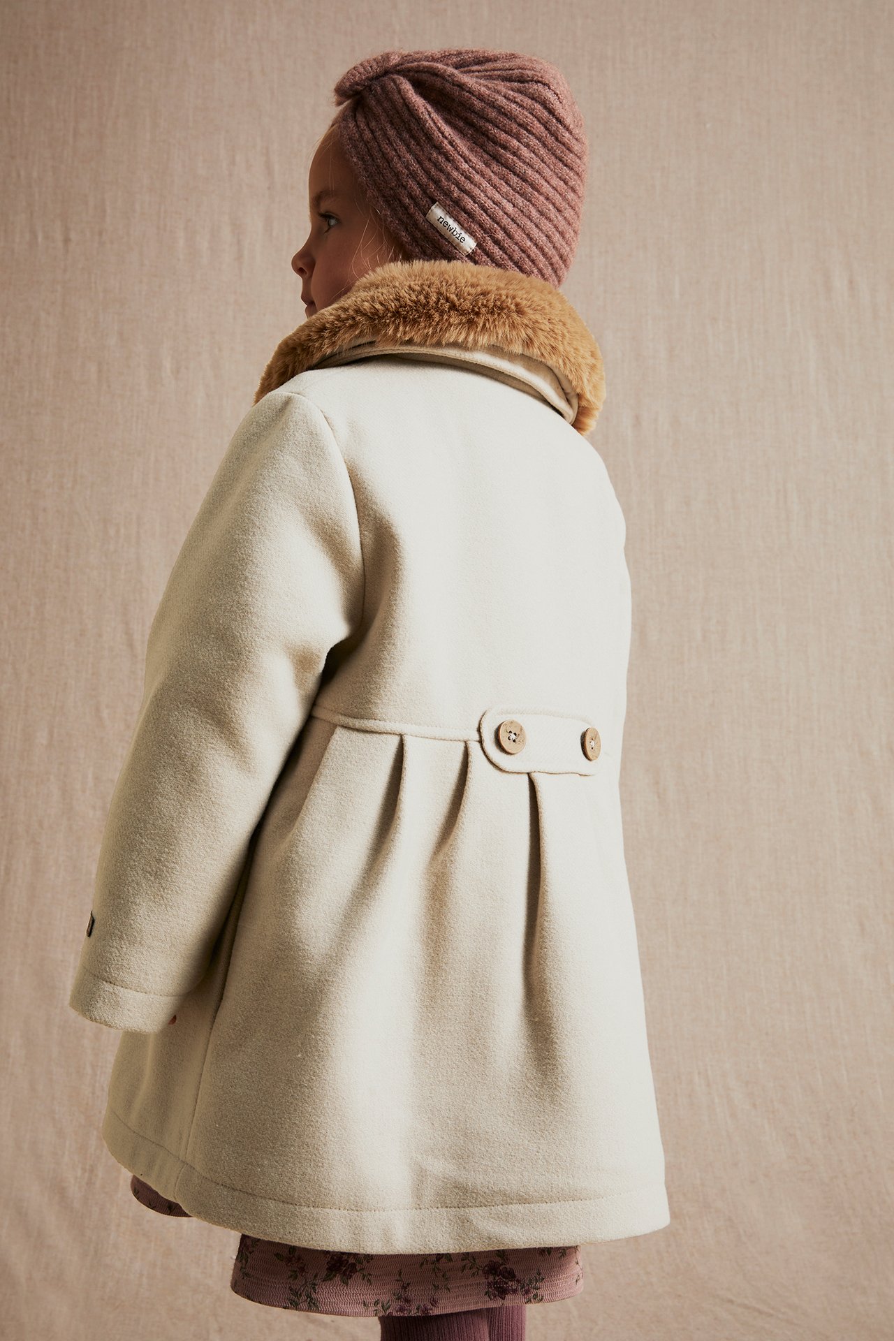 Back view of a kid in a beige woven coat with a brown faux fur collar, decorative back buttons, and a pink ribbed turban.