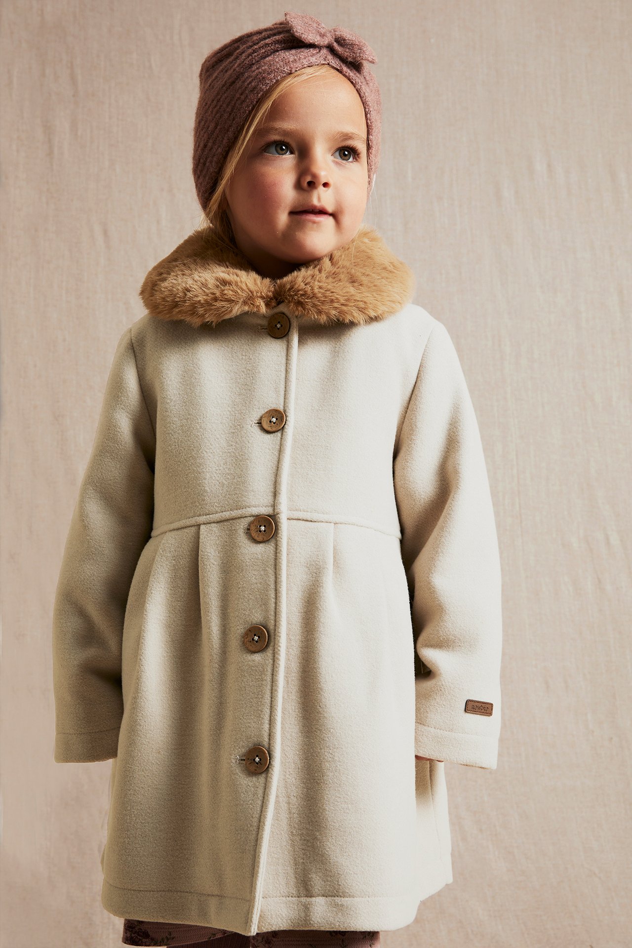 Front view: Kid in a beige woven coat with brown faux fur collar, coconut buttons, and pink ribbed turban.