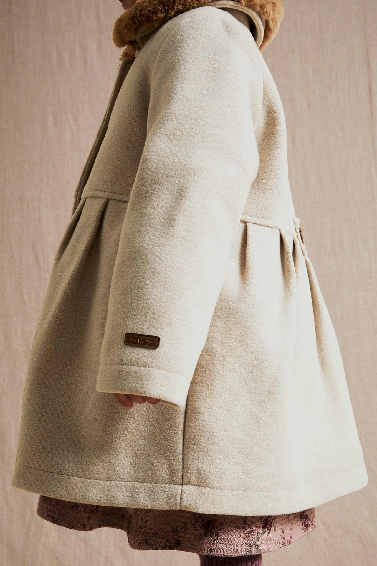 Side view of a kid wearing a beige woven coat with a brown faux fur collar over a pink floral dress.
