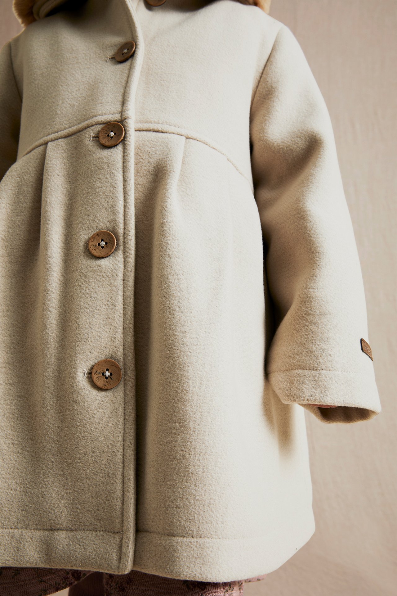 Close-up front view of a kid's beige woven coat with brown buttons and a faux fur collar.