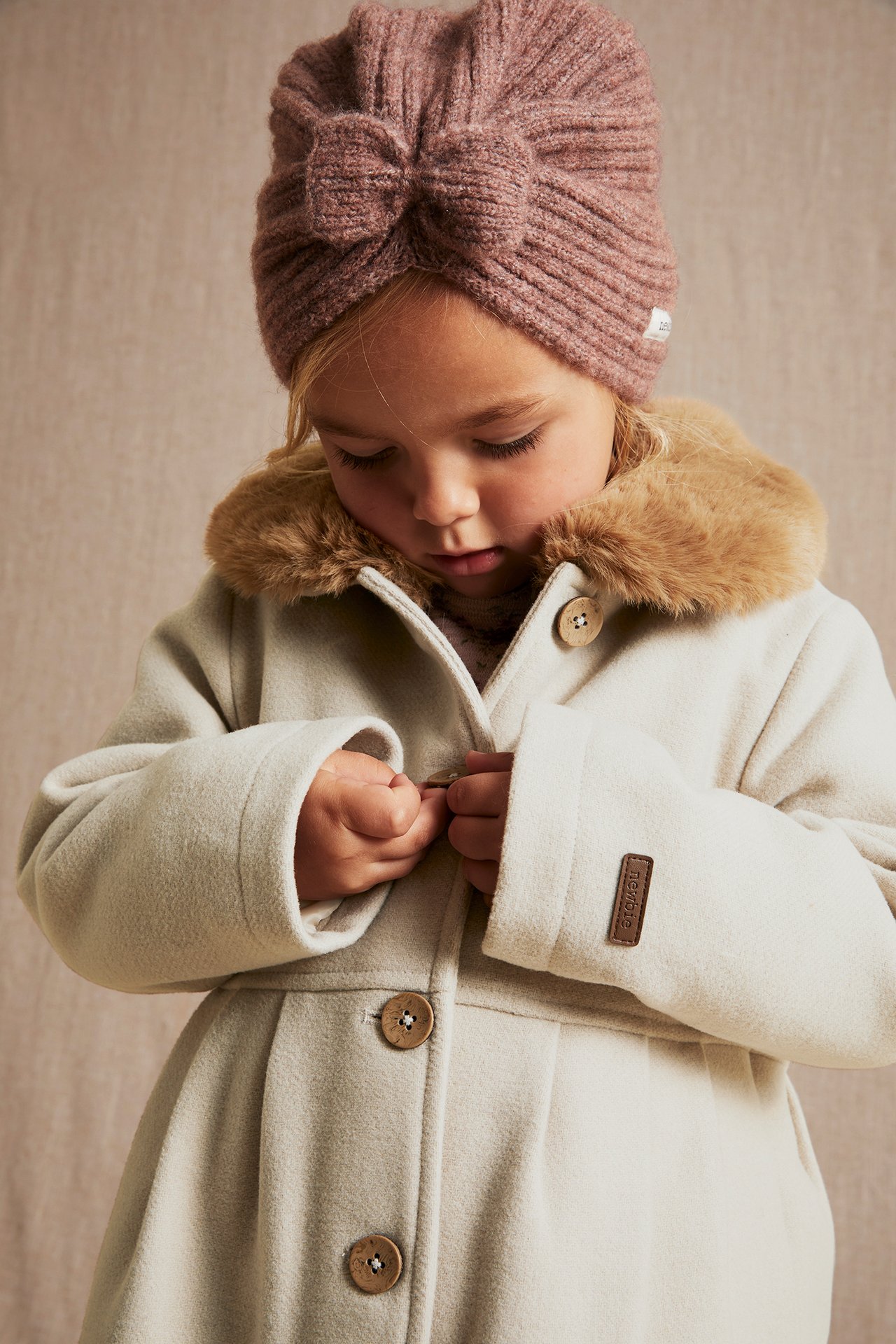 Close-up of a kid buttoning a beige woven coat with a brown faux fur collar, wearing a pink knitted turban.