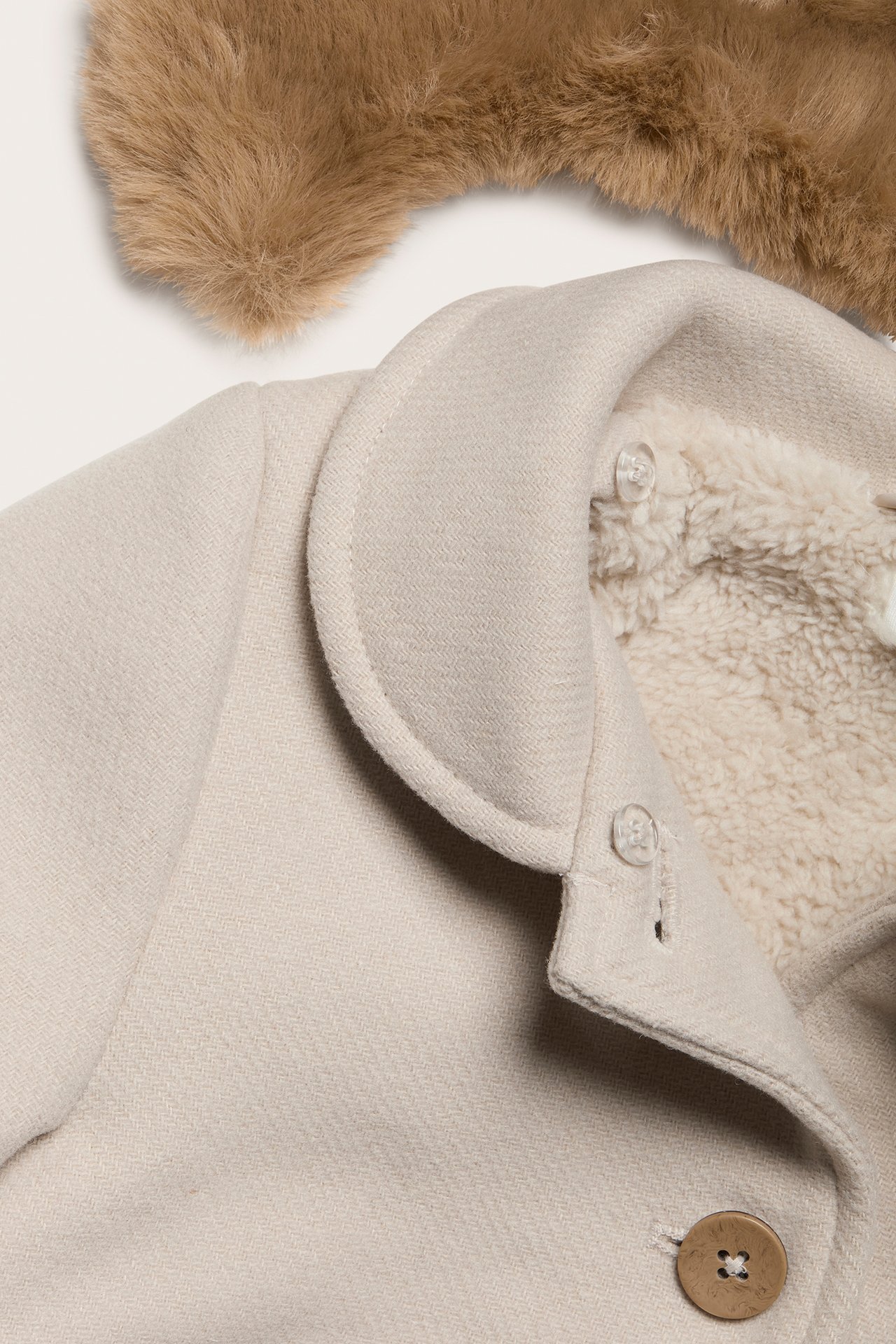 Close-up of a beige woven coat with pile lining, coconut buttons, and a brown faux fur collar.