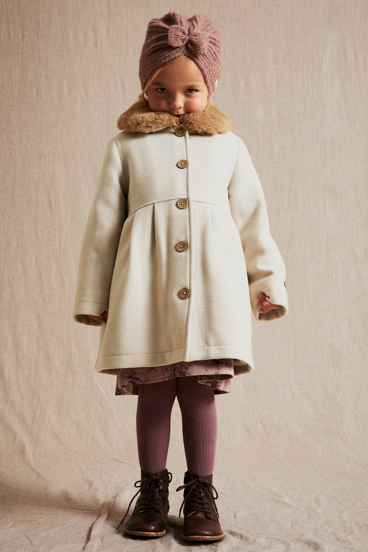 Front view of a kid in a beige coat with faux fur collar, pink turban, floral dress, purple tights, brown boots.