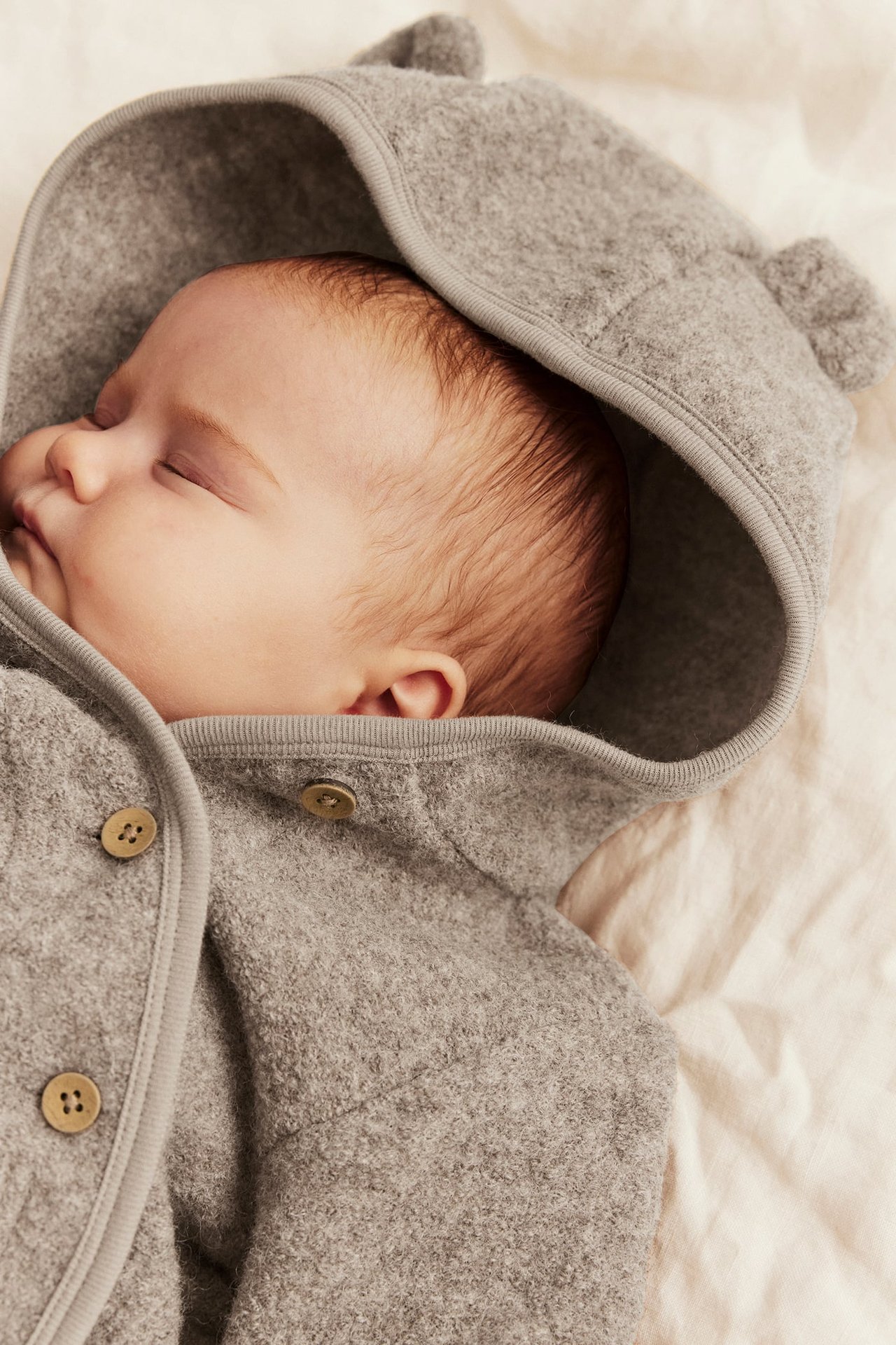 Nahaufnahme eines schlafenden Babys in einer grauen Filz-Kapuzenjacke mit Bärenohren und Knöpfen.