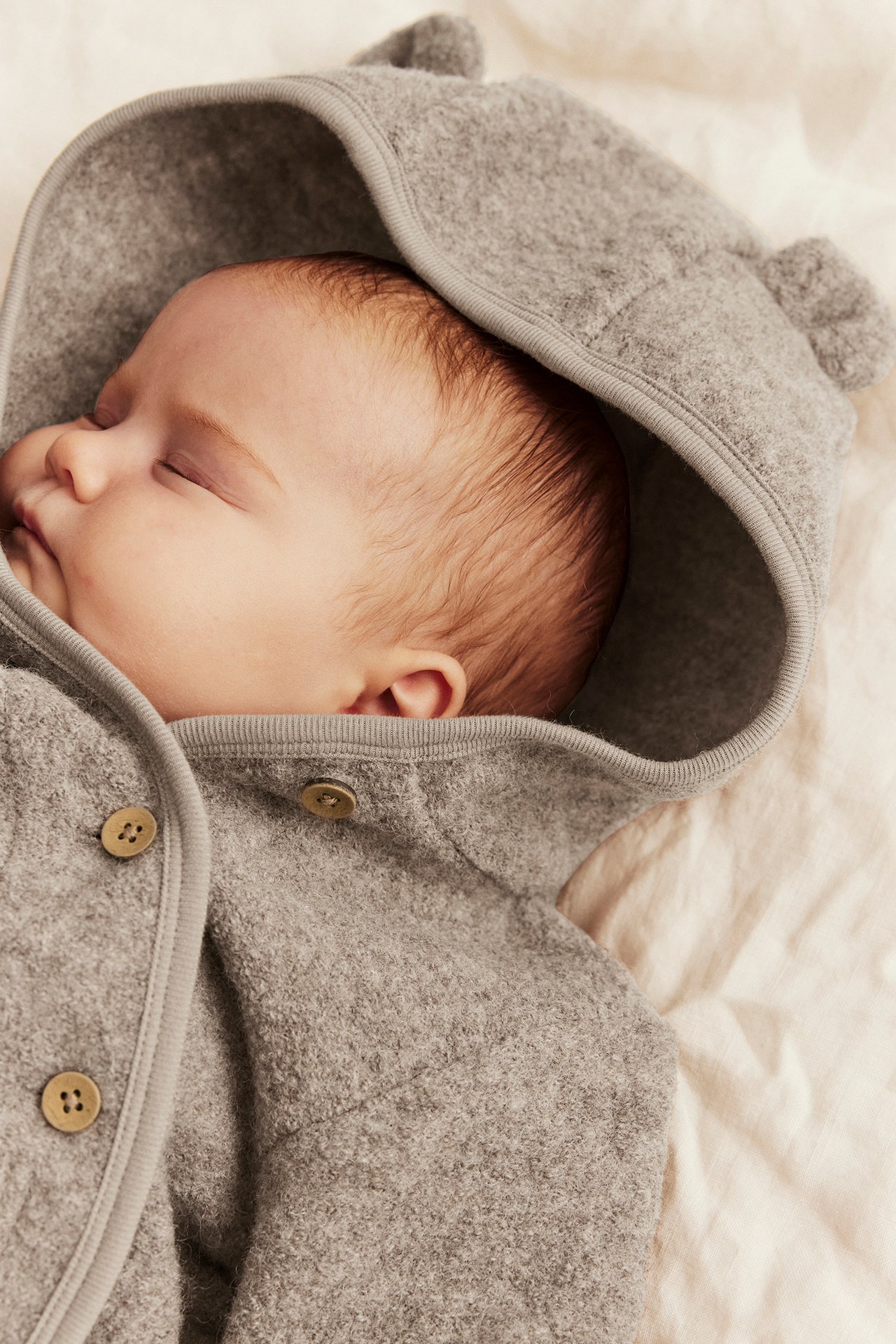 Close-up of a sleeping baby in a grey felt hooded jacket with bear ears and buttons.