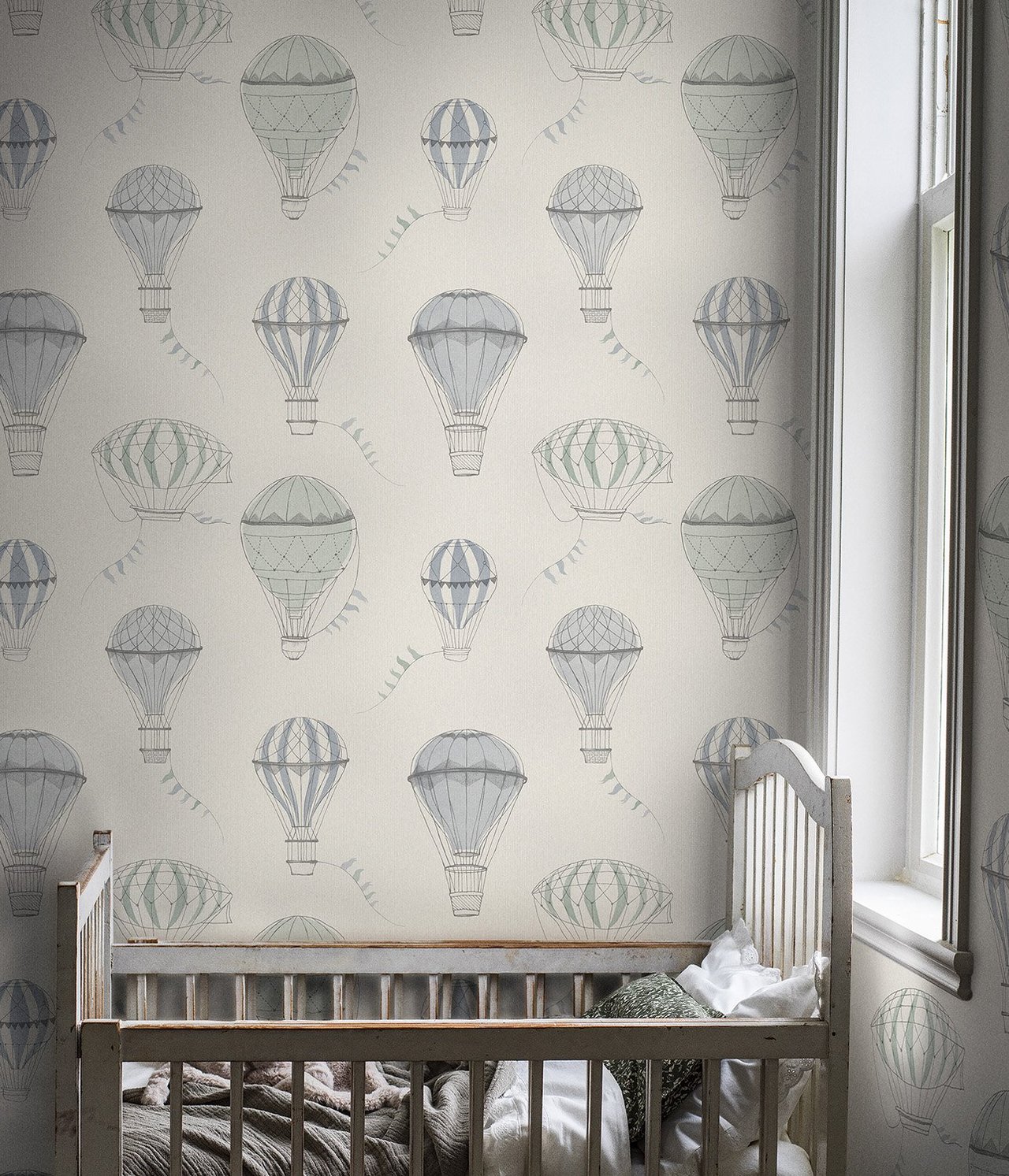 Vorderansicht: grau-beige Tapete mit blauen und grünen Heißluftballons, ein weißes Holzbettchen.