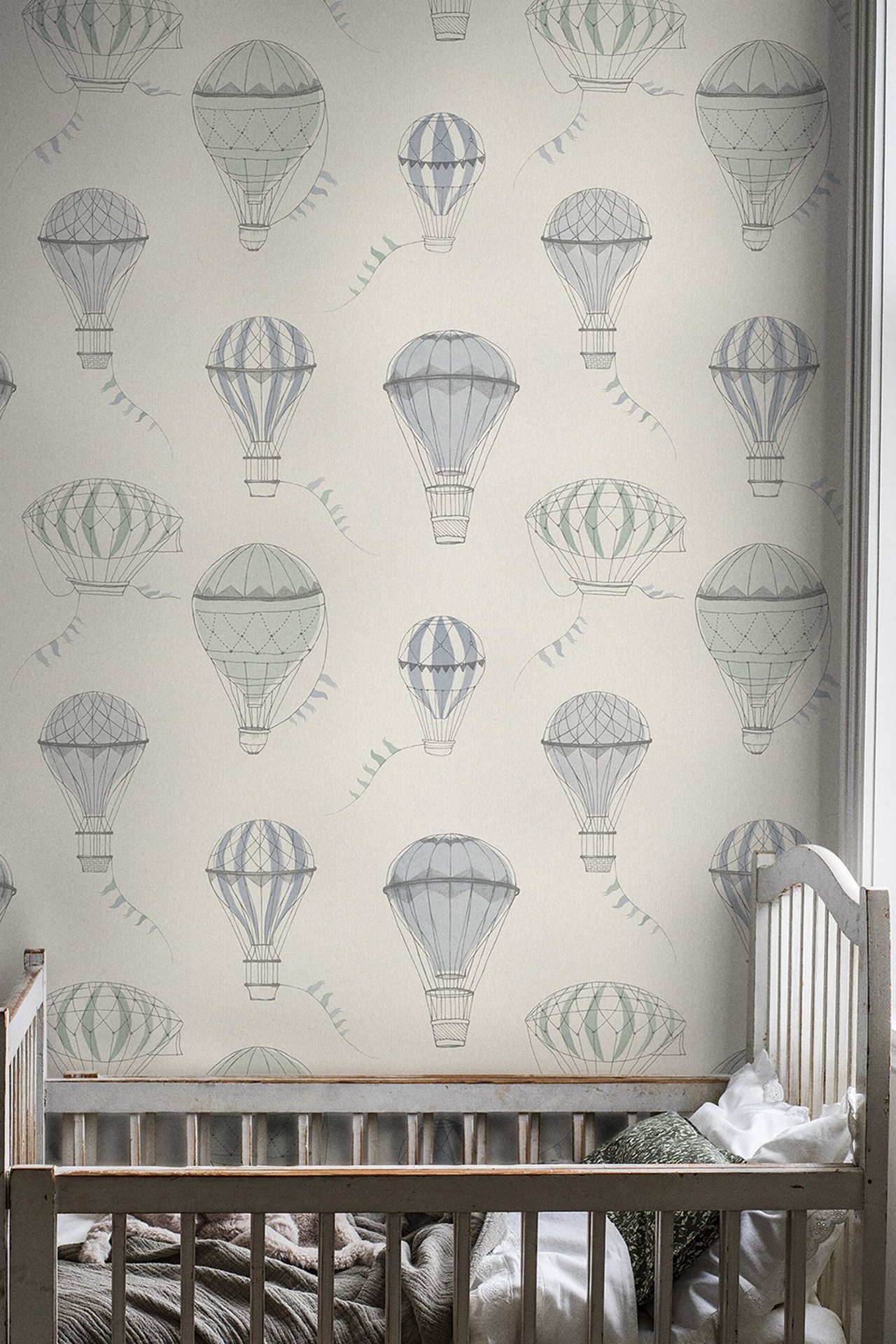 Front view of grey-beige wallpaper with blue and green pastel hot air balloons, behind a white baby crib.