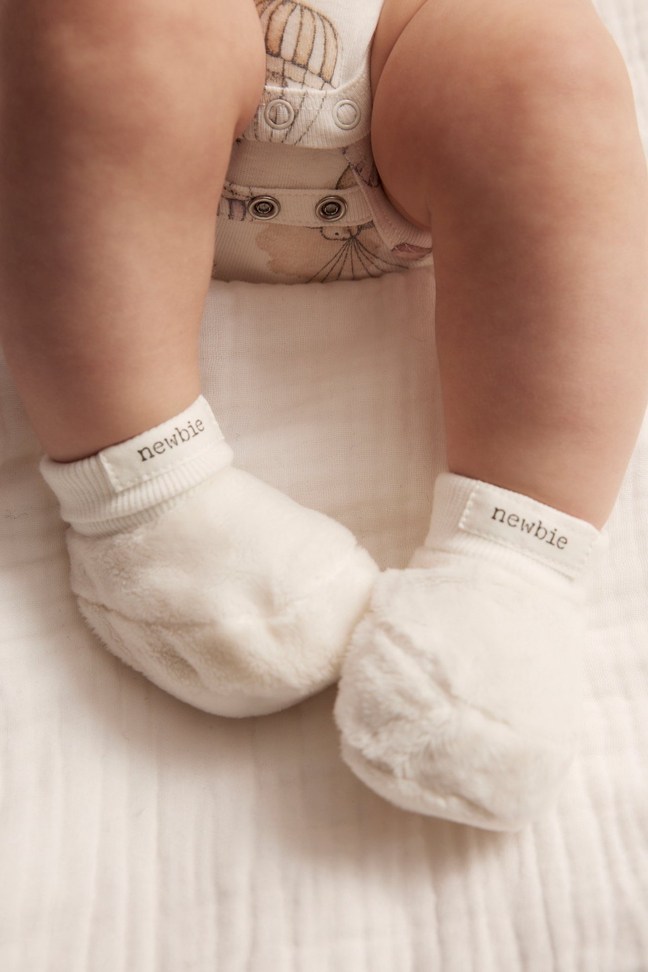 Close-up of a baby's feet wearing off-white fluffy booties with 'newbie' labels, over a patterned bodysuit.