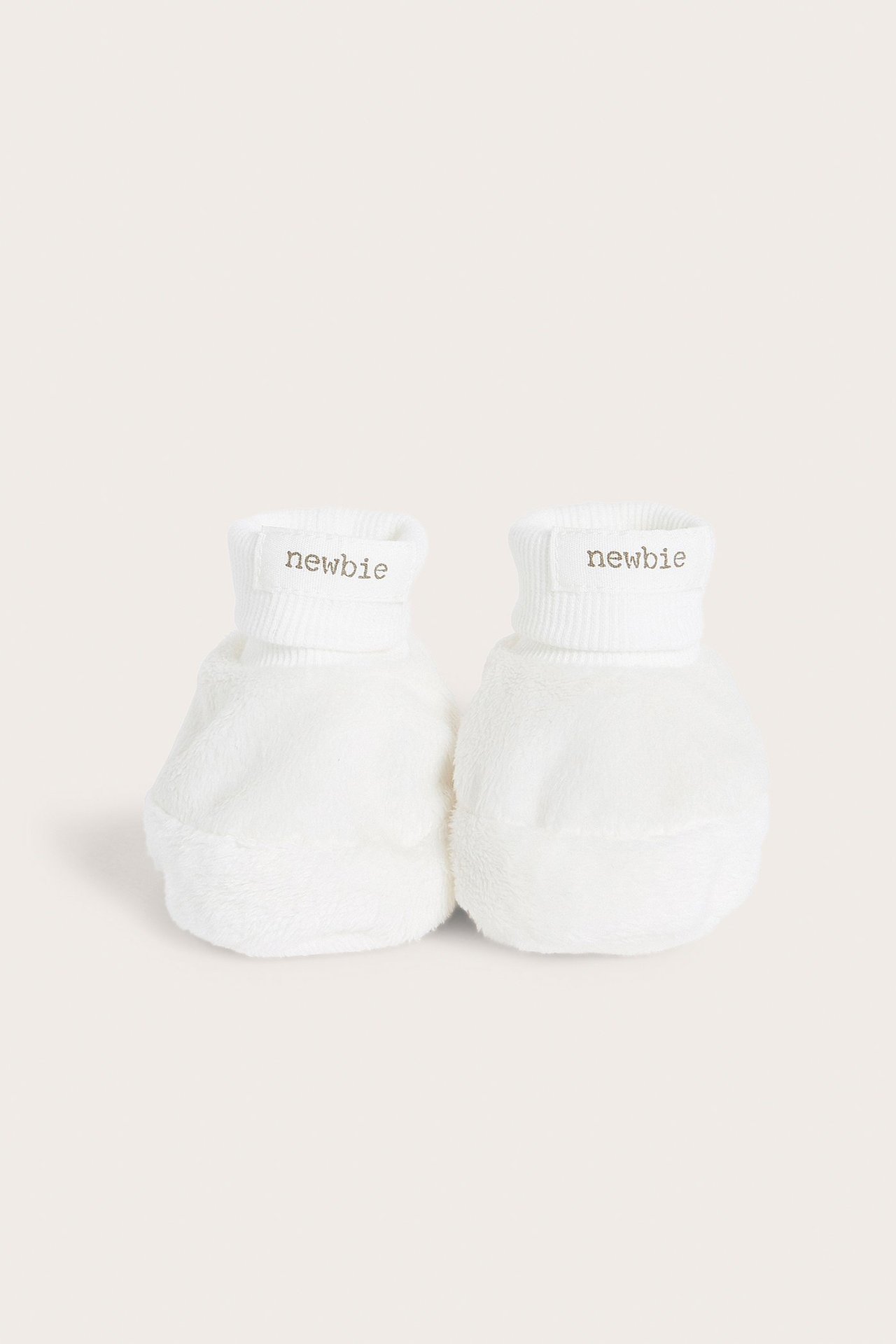 Vorderansicht eines Paares cremeweißer flauschiger Babyschuhe mit gerippten Bündchen und 'newbie'-Etiketten.