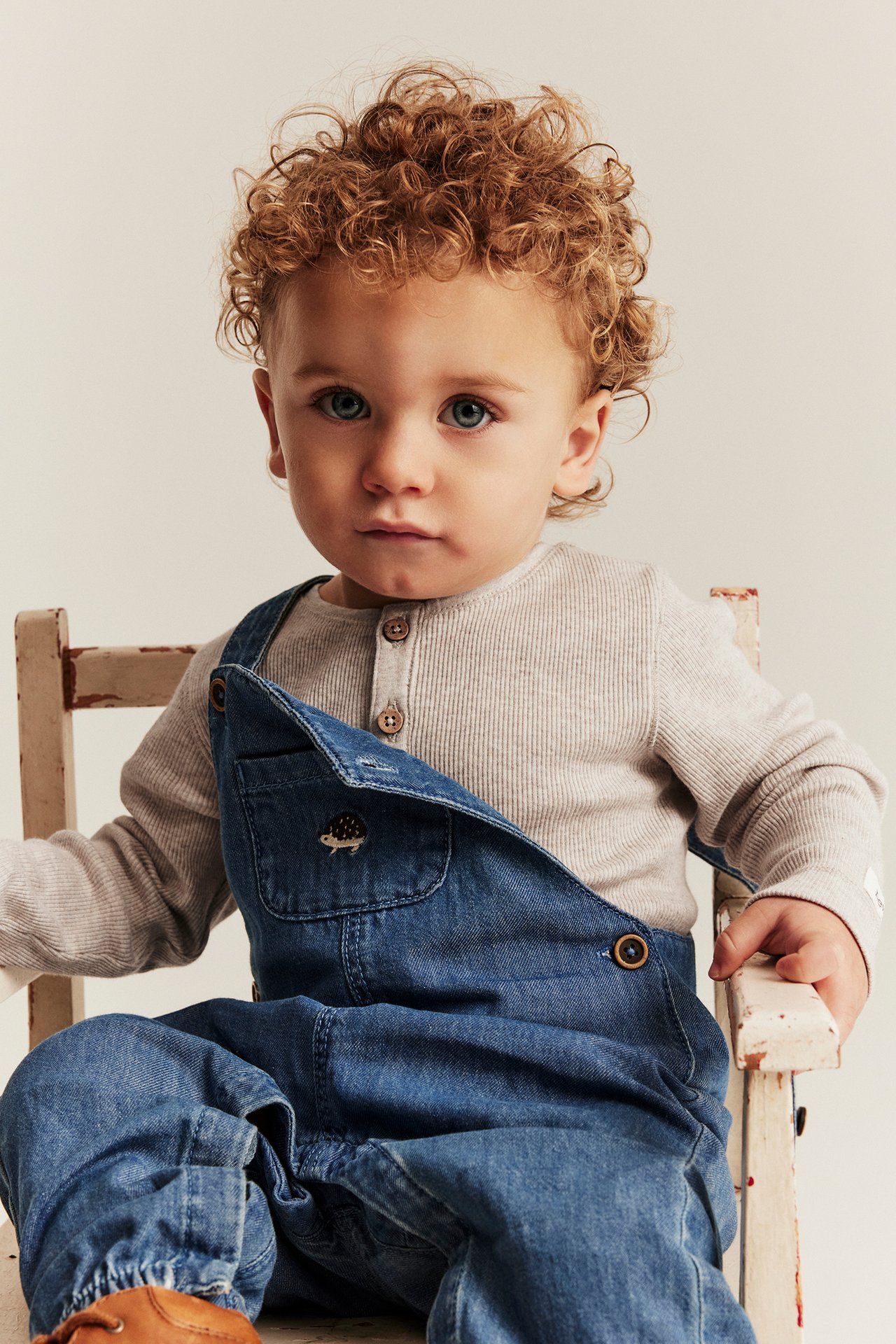 Front view of a baby wearing a beige melange ribbed long-sleeved bodysuit under blue denim overalls.