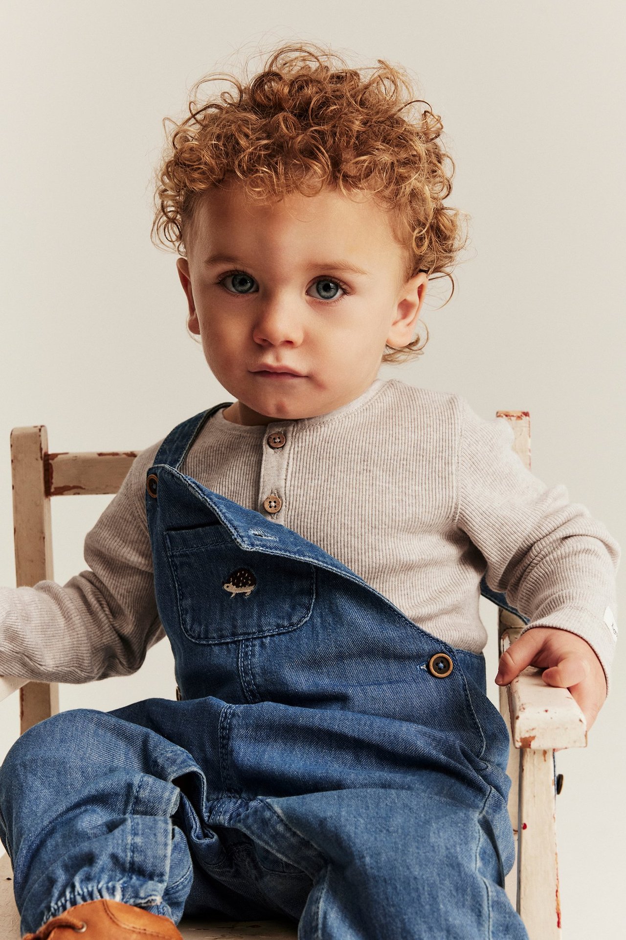 Front view of a baby wearing a beige melange ribbed long-sleeved bodysuit under blue denim overalls.