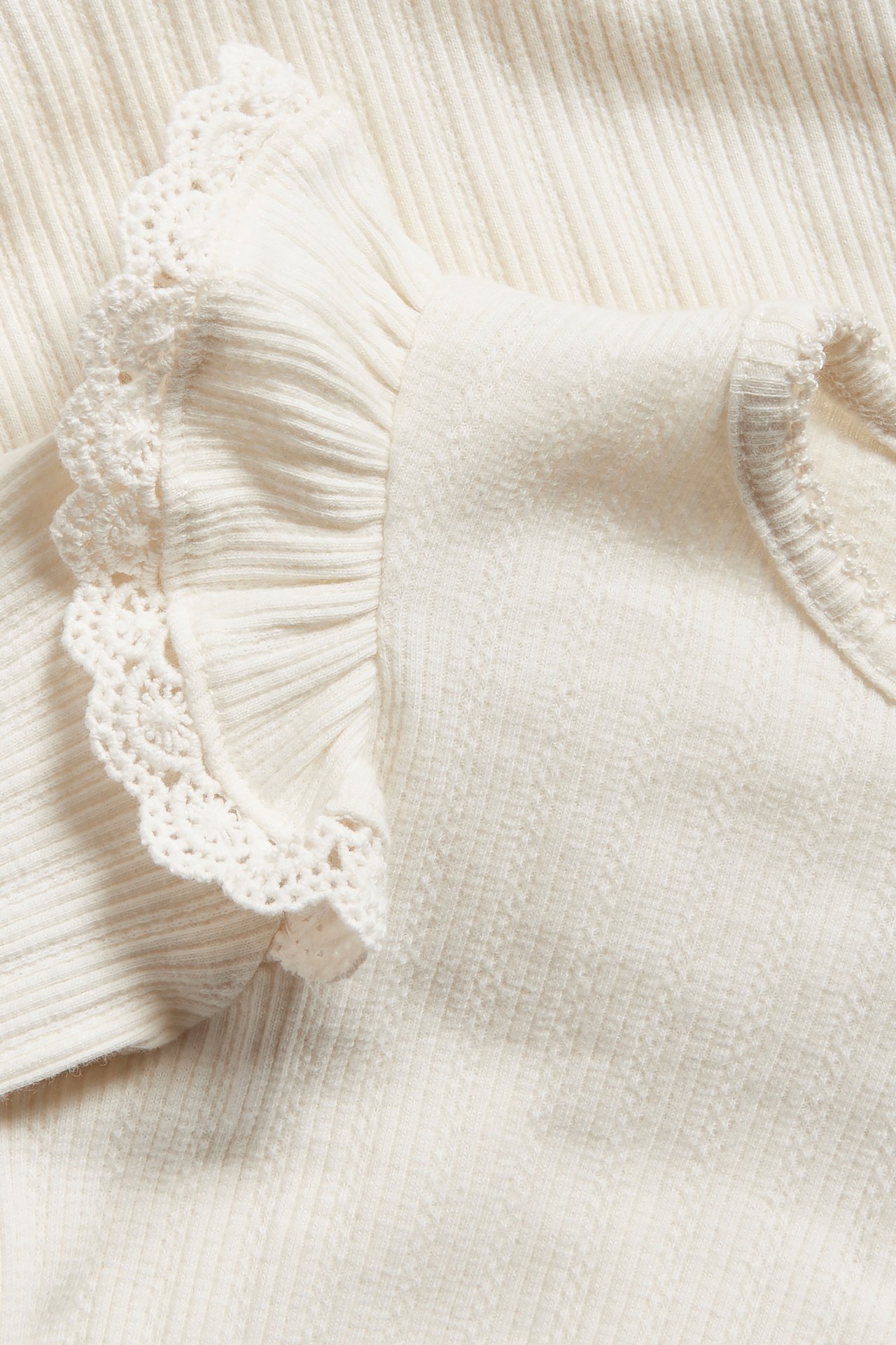 Nahaufnahme eines cremefarbenen gerippten Baby-Bodys mit langen Ärmeln, Rüschenschultern und Spitzenbesatz.