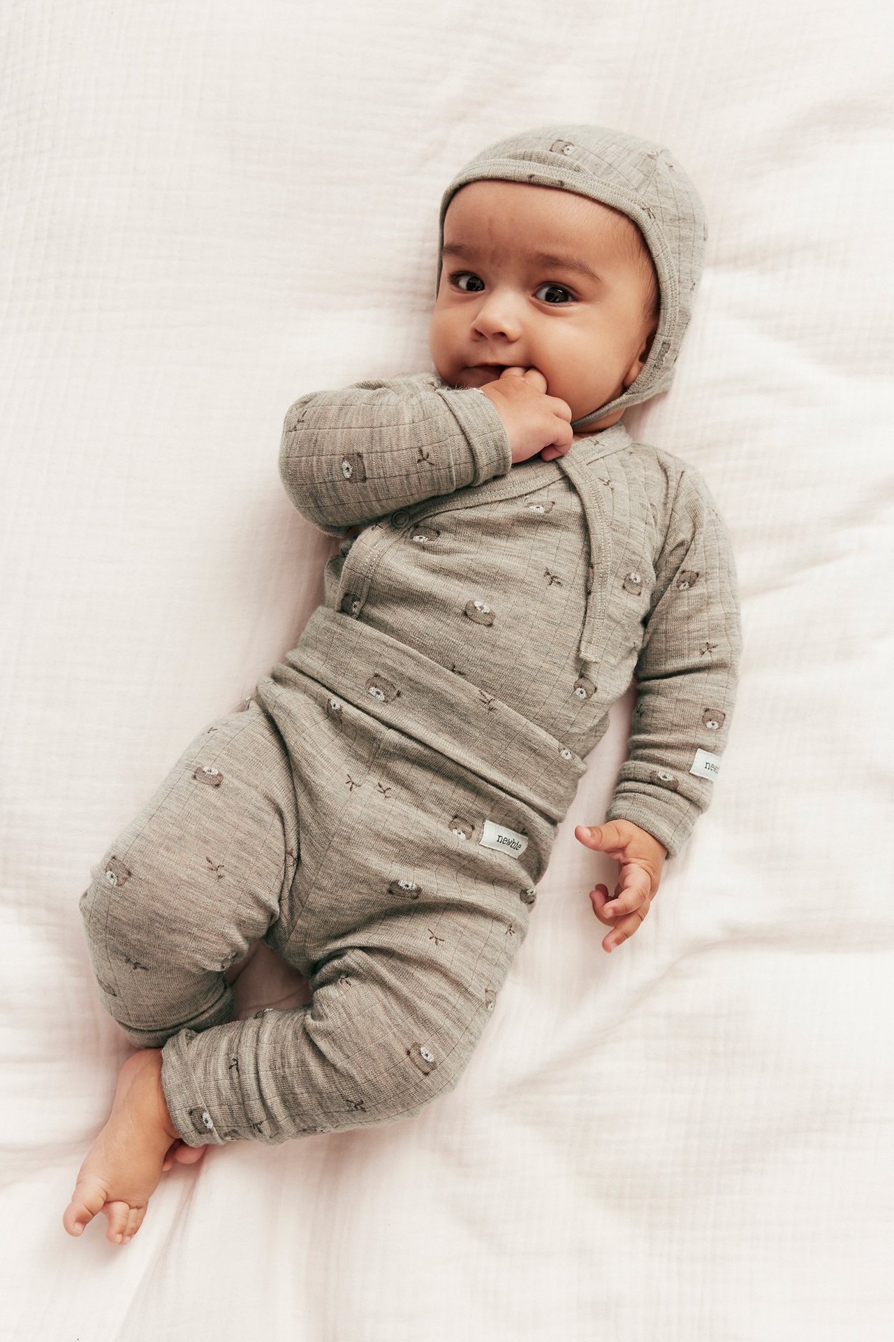 Draufsicht eines Babys in beigemelierter gerippter Woll-Leggings mit Teddybärmuster, passendem Wickeloberteil und Mütze.