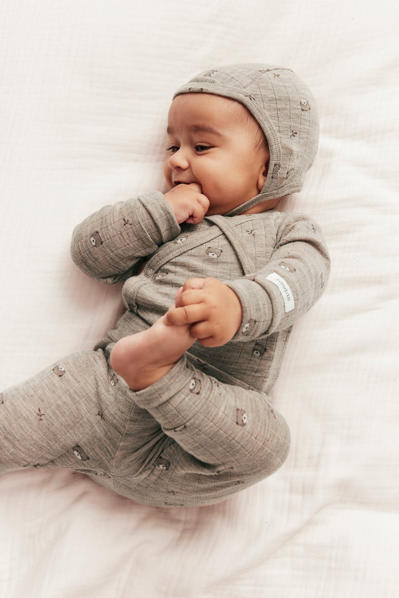 Baby trägt einen beige melierten gerippten Wollbody mit Teddybärmuster und passender Mütze.