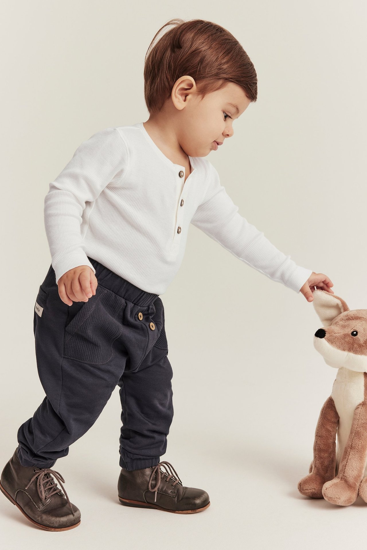 Front view: baby boy in white ribbed long-sleeved body with buttons, dark trousers, brown boots, touching fox toy.