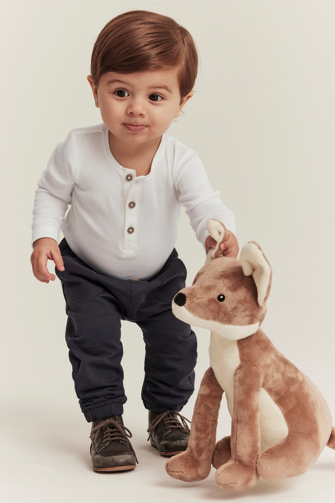Vorderansicht: Baby in weißem geripptem Langarmbody, dunkler Hose, braunen Schuhen, hält Plüschfuchs.