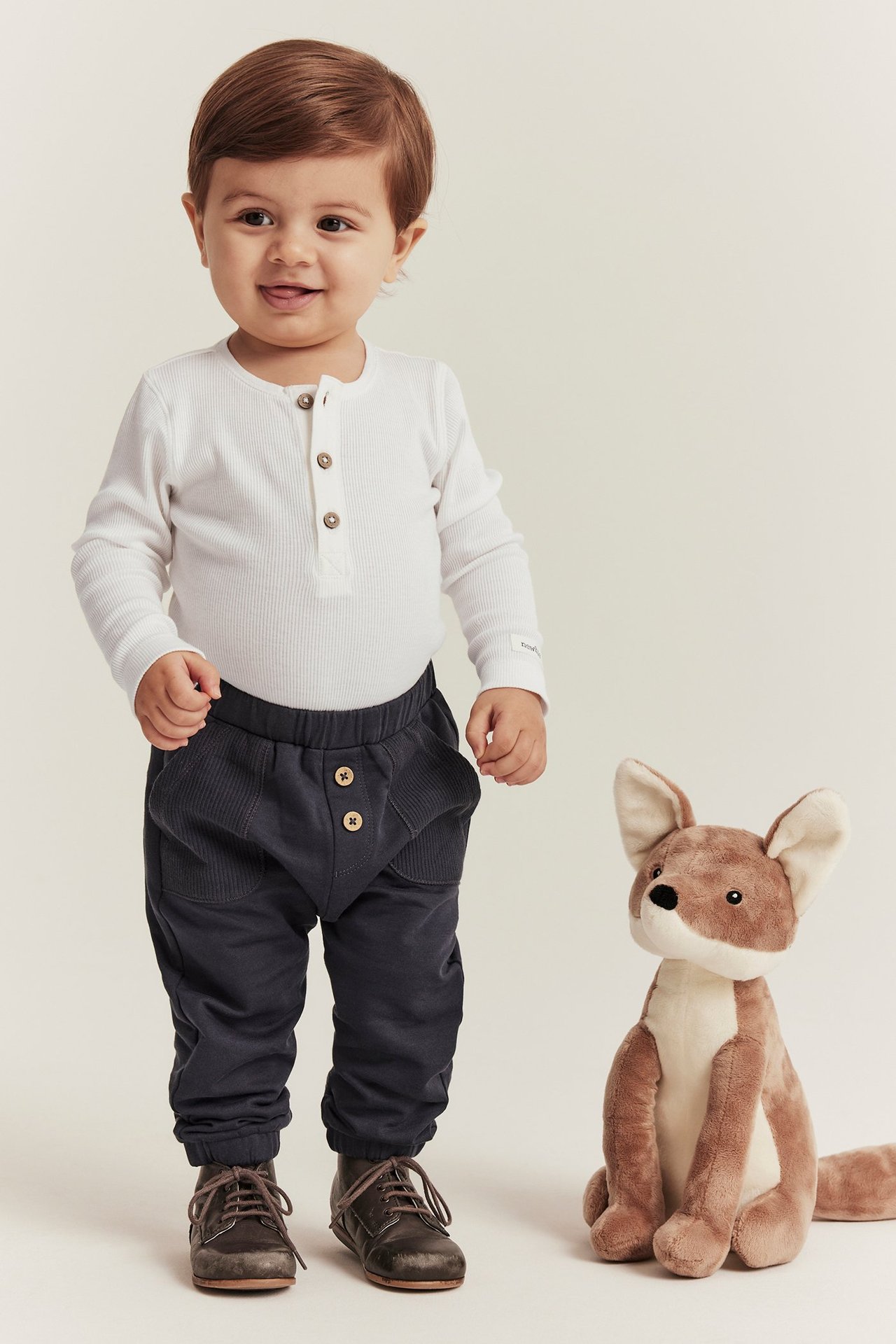 Front view: Baby in white ribbed long-sleeved body, dark grey trousers, brown shoes, with a plush fox.