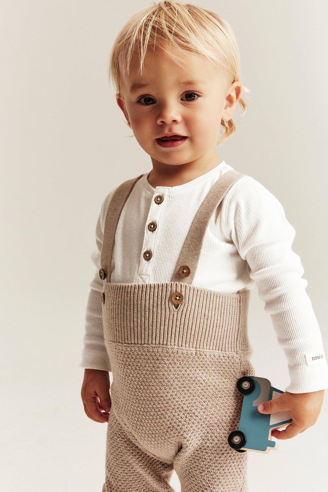 Baby in weißem geripptem Langarmbody mit Knöpfen, unter beiger Strick-Latzhose, hält Spielzeugauto.