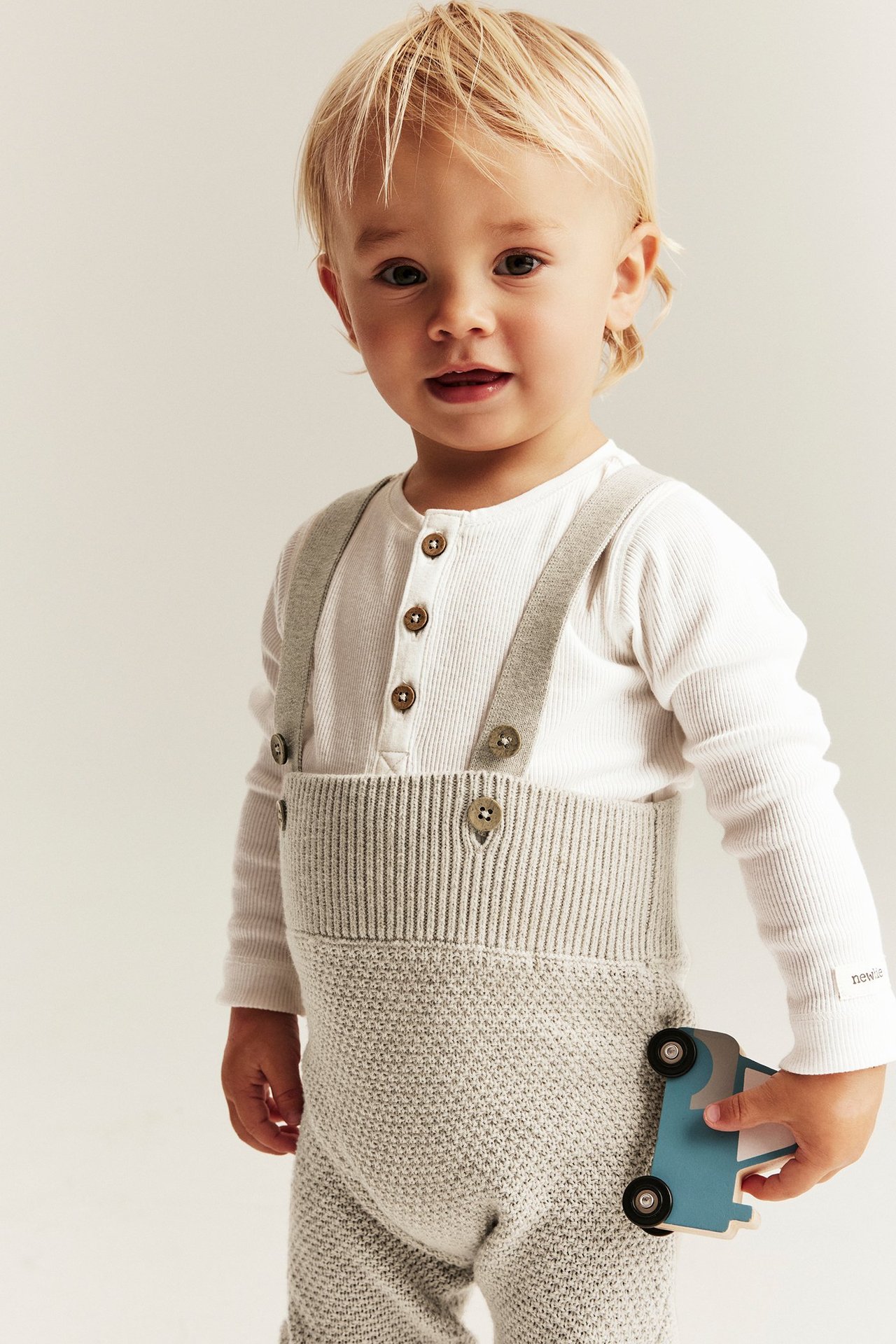 Front view, baby in white ribbed long-sleeved body and light grey knitted dungarees, holding a toy car.