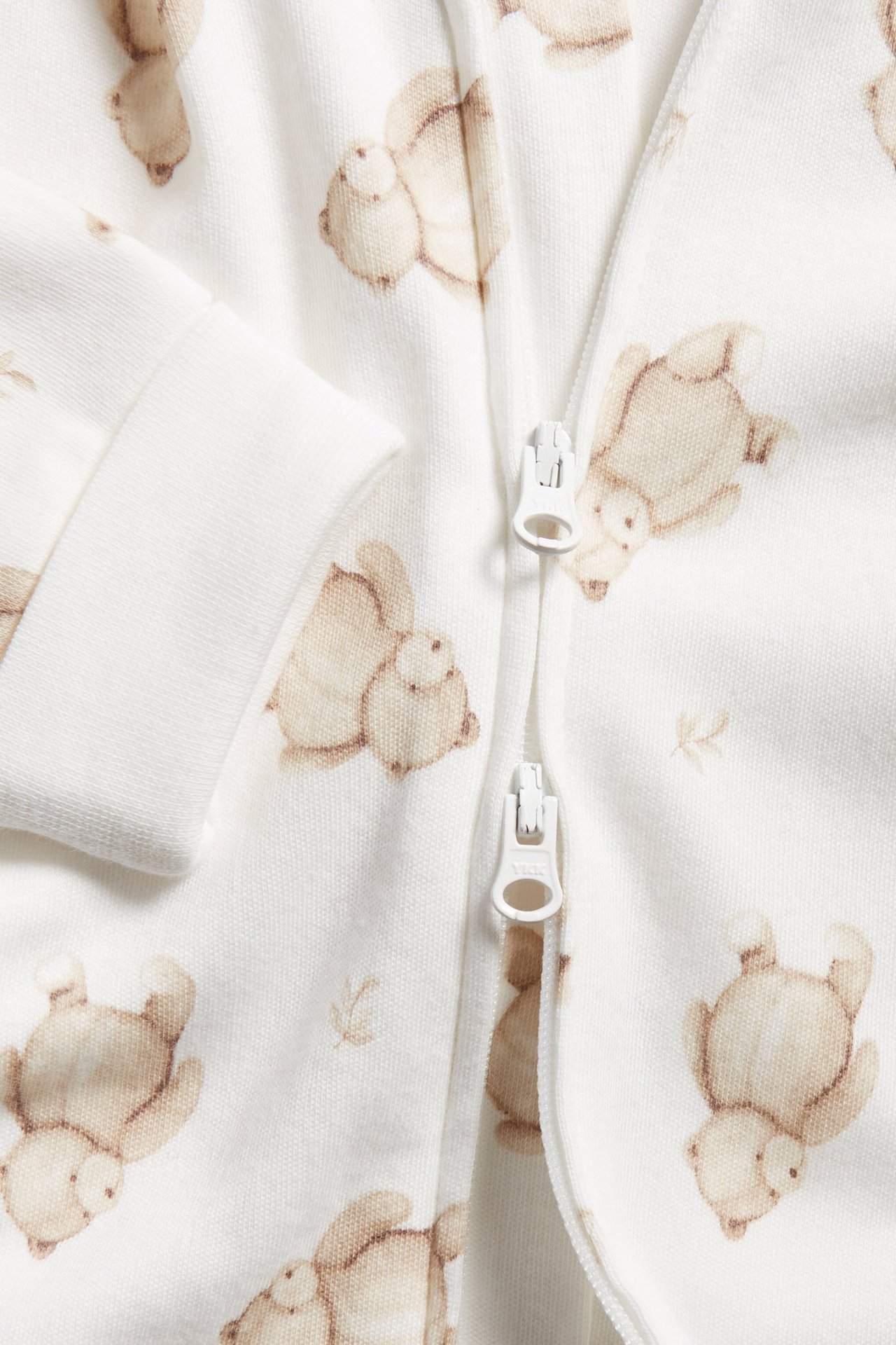 Close-up of an off-white baby sleepsuit with a brown teddy bear pattern and a white two-way zipper.
