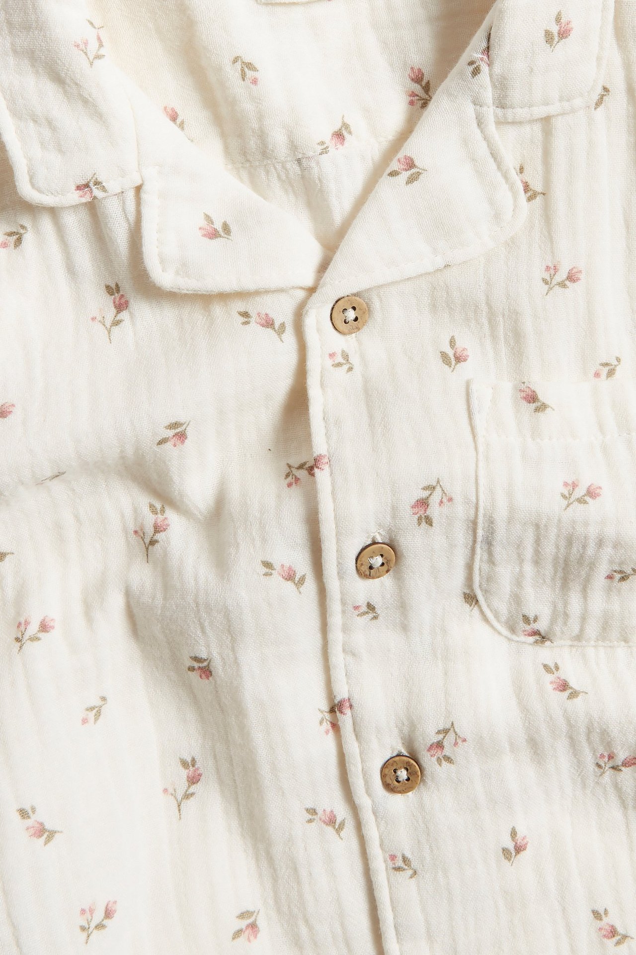 Close-up front view: beige cotton short-sleeve shirt with small pink floral print, collar, wooden buttons, chest pocket.