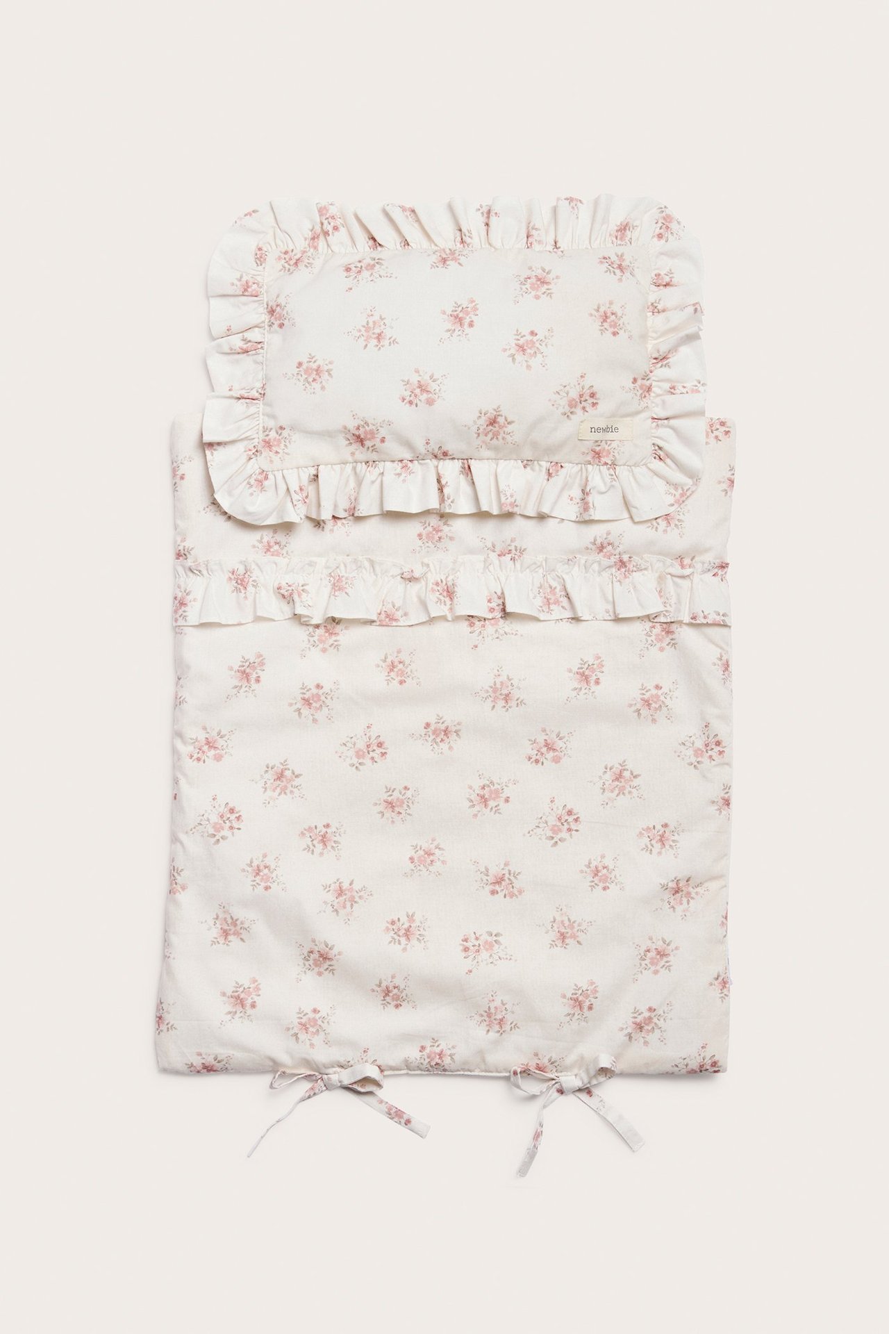 Draufsicht auf ein cremefarbenes Puppenbettwäsche-Set mit rosa Blumenmuster, Rüschen und Bindebändern.