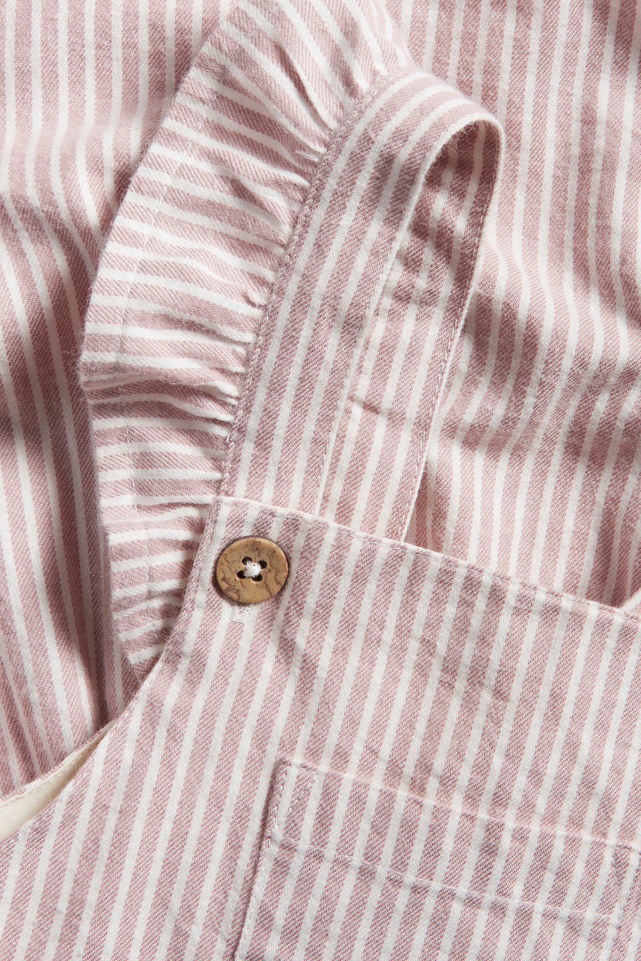 Nahaufnahme eines rosa-weiß gestreiften Latzkleides mit Rüschen an den verstellbaren Trägern und Holzknopf.