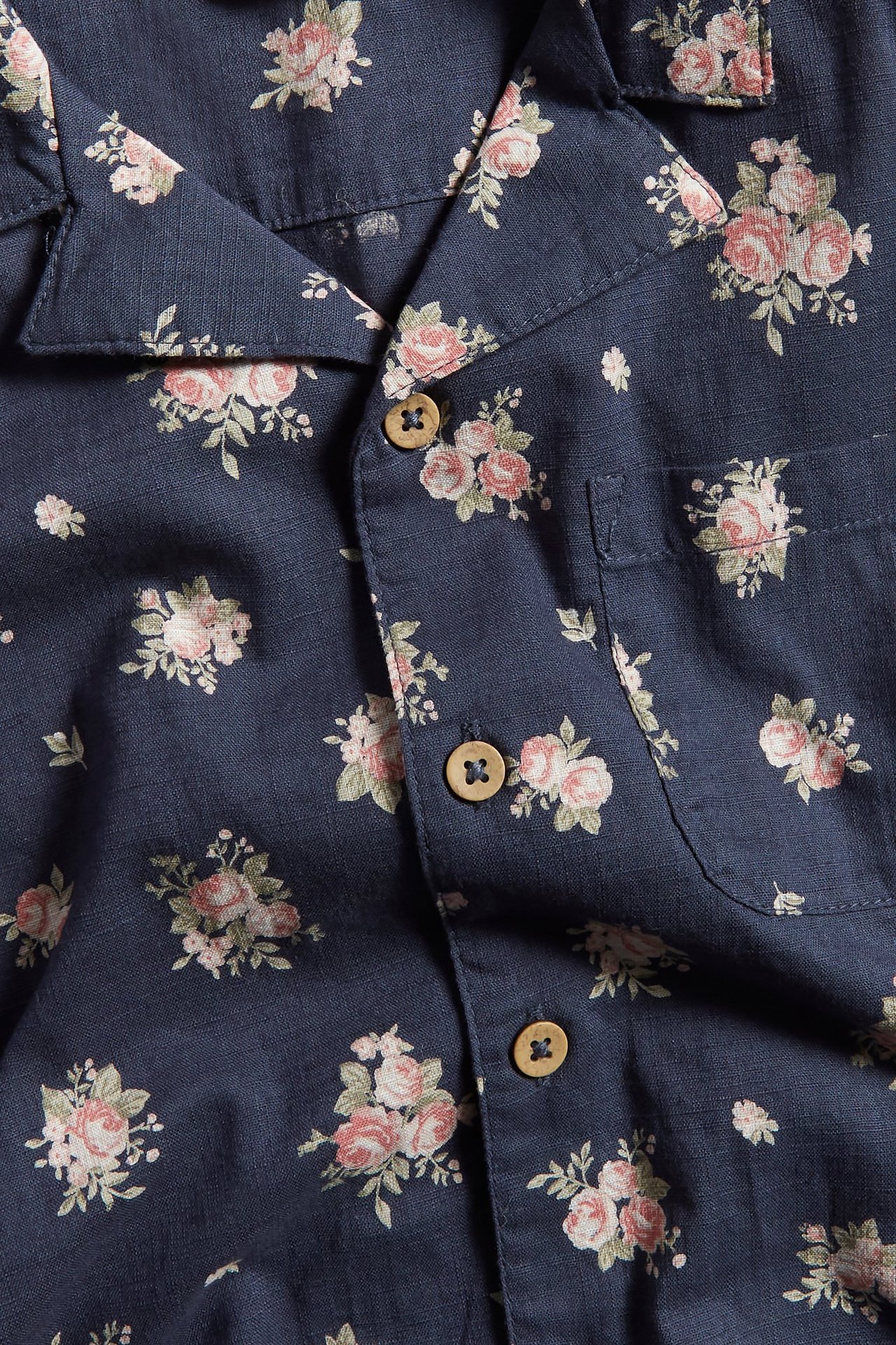 Nahaufnahme von einem dunkelblauen kurzärmeligen Baumwollhemd mit rosa Blumenmuster und Holzknöpfen.