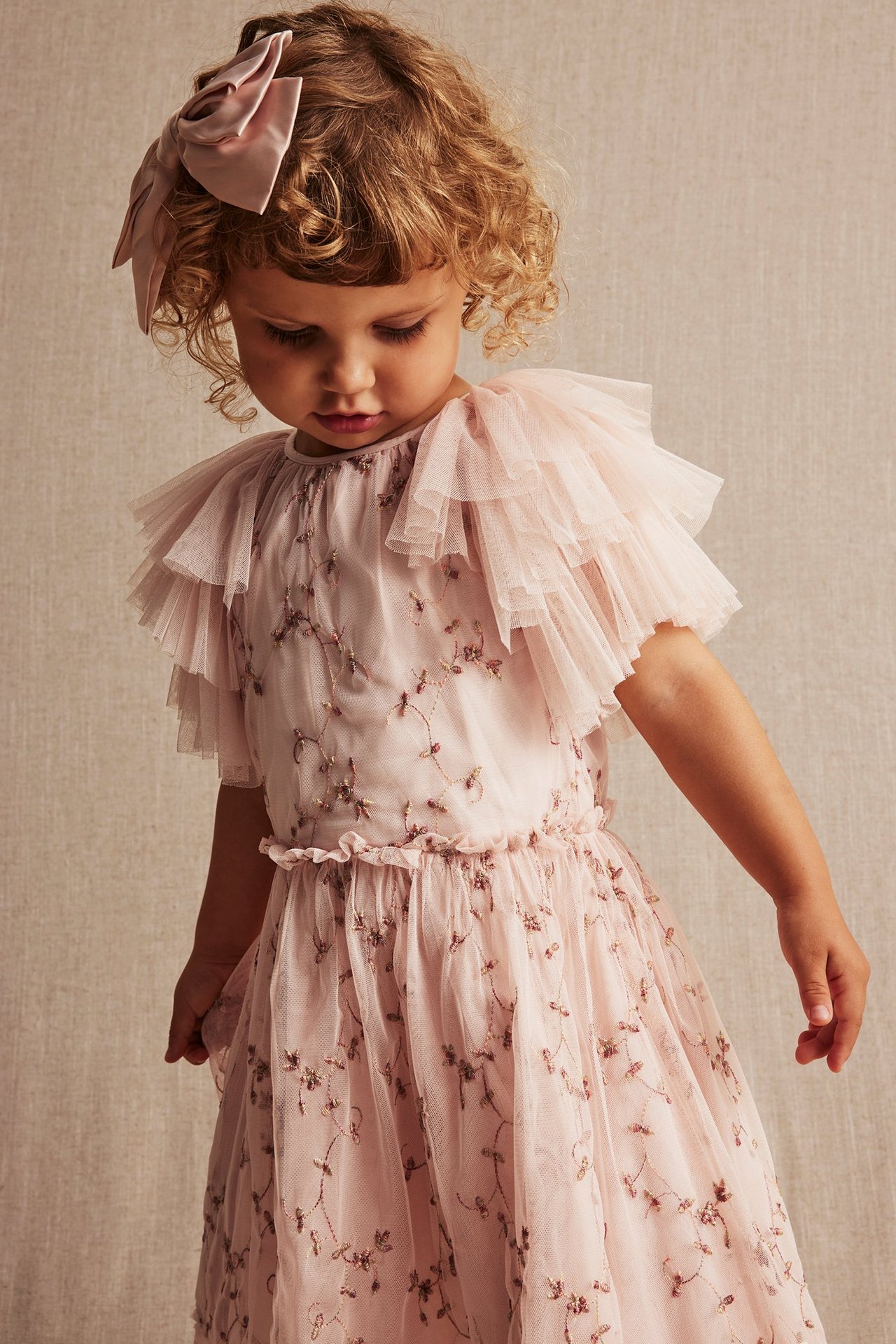 Vorderansicht: Baby in einem hellrosa, blumig bestickten Netz-Kleid mit Rüschenärmeln und rosa Haarschleife.