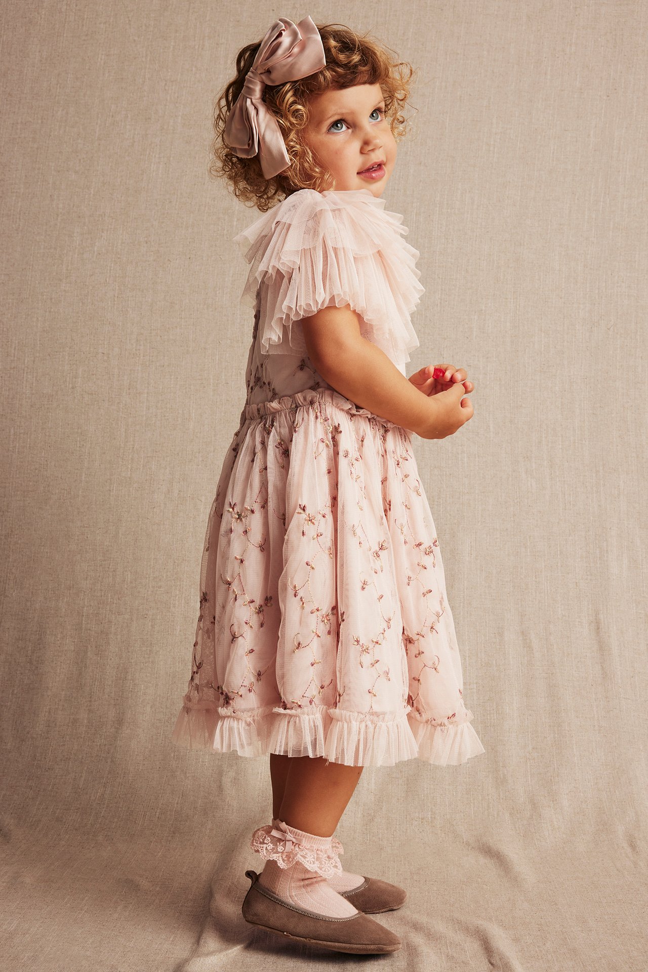 Seitenansicht eines Kindes in hellrosa besticktem Netz-Kleid mit Rüschenärmeln, rosa Schleife, Rüschensocken und braunen Schuhen.