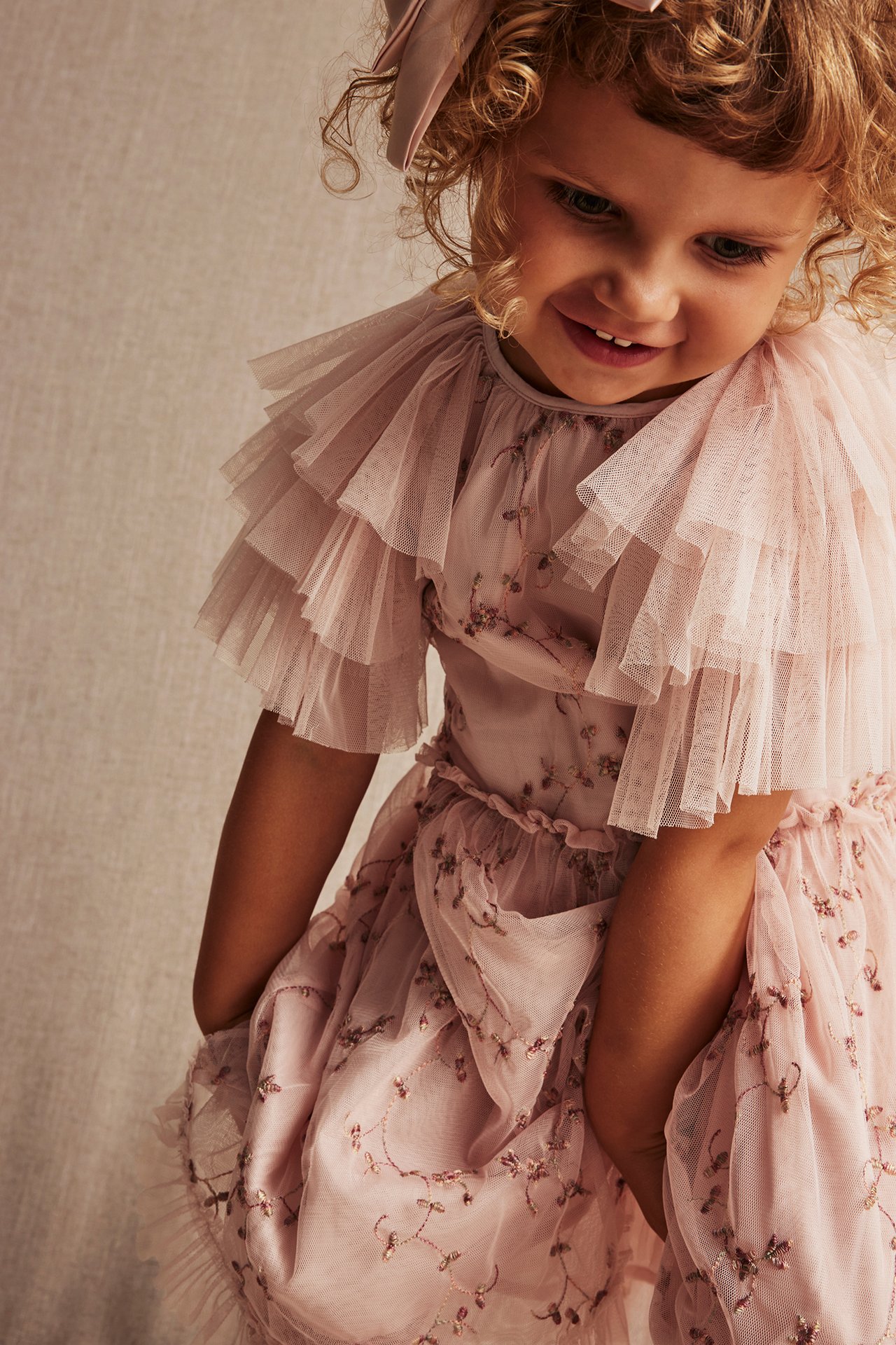 Nahaufnahme eines Kindes in einem hellrosa bestickten Netz-Kleid mit Rüschenärmeln und einer rosa Haarschleife.