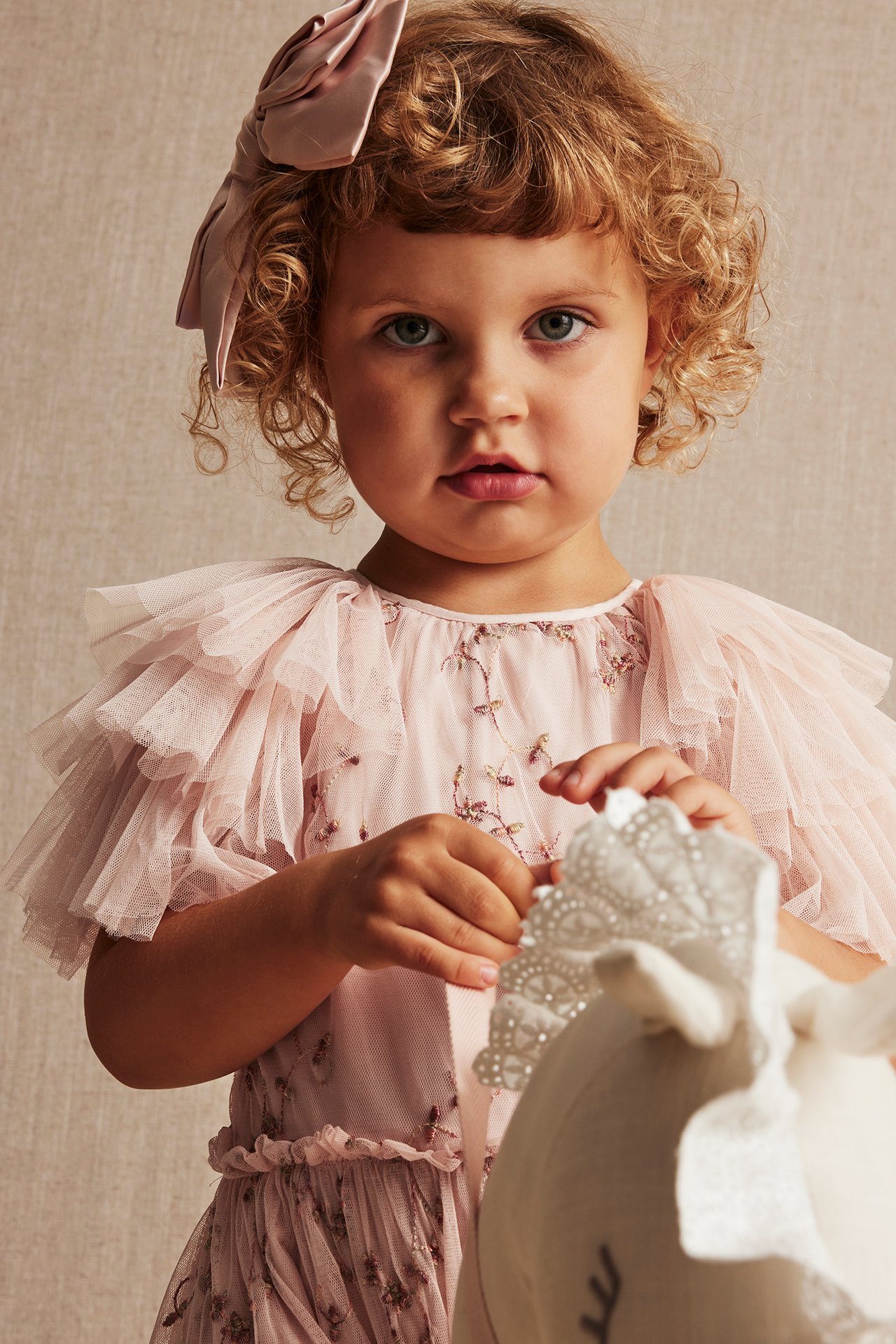 Vorderansicht: Kind in hellrosa besticktem Netz-Kleid mit Rüschenärmeln, rosa Schleife, hält weißes Spielzeug.