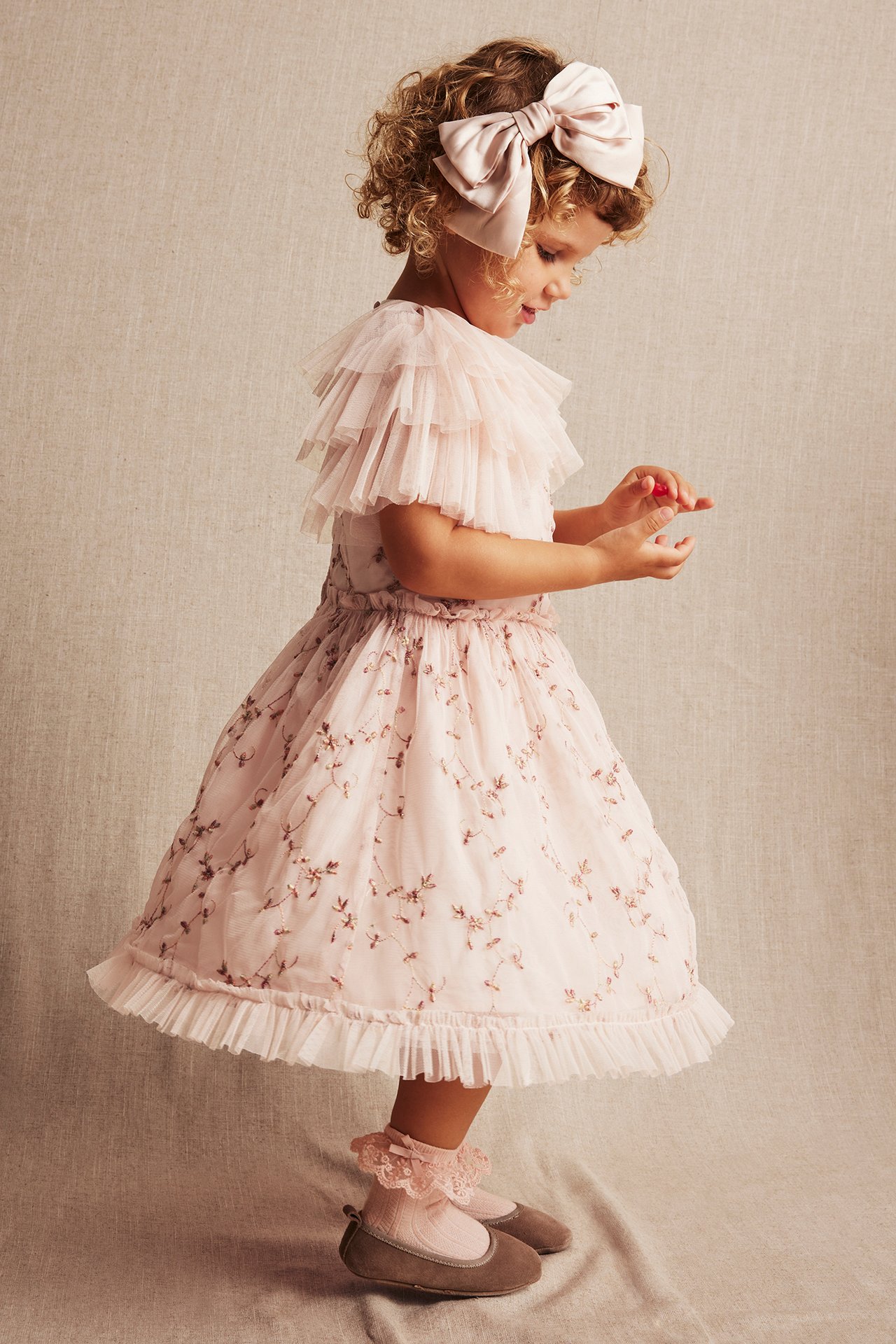 Kind in einem hellrosa blumig bestickten Netz-Kleid, rosa Schleifen-Haarband, Spitzensocken und braunen Ballerinas.