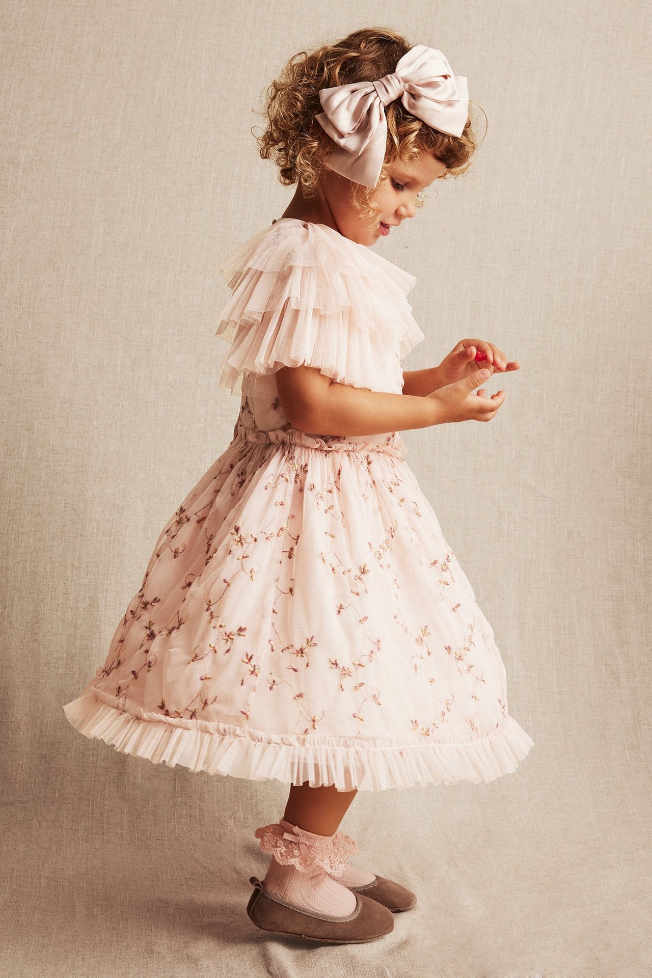 Seitenansicht eines Kindes in einem hellrosa bestickten Netz-Kleid, rosa Schleife, rosa Spitzensocken und braunen Ballerinas.