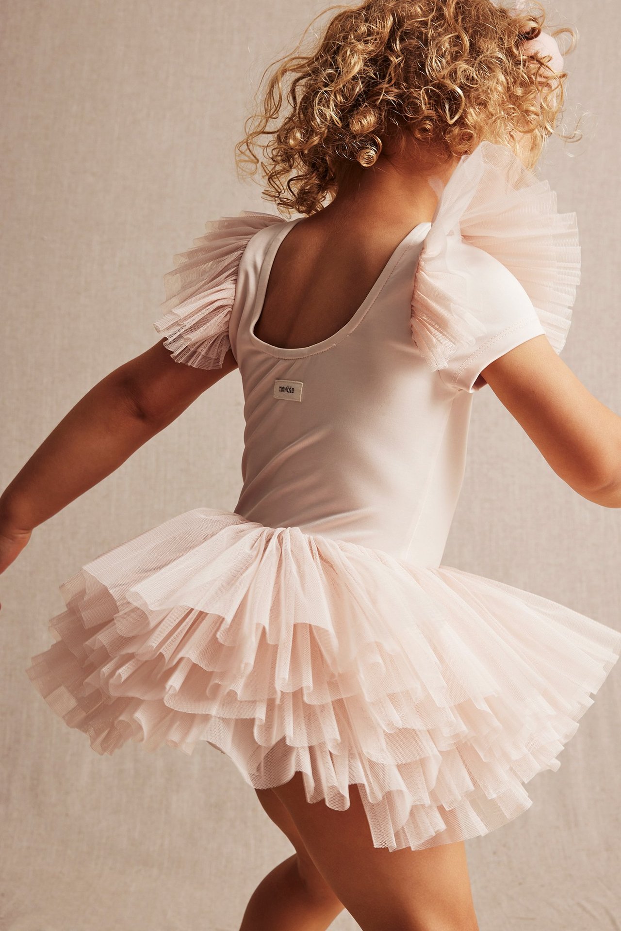 Back view of a kid wearing a light pink ballerina leotard with layered mesh ruffles on sleeves and skirt.