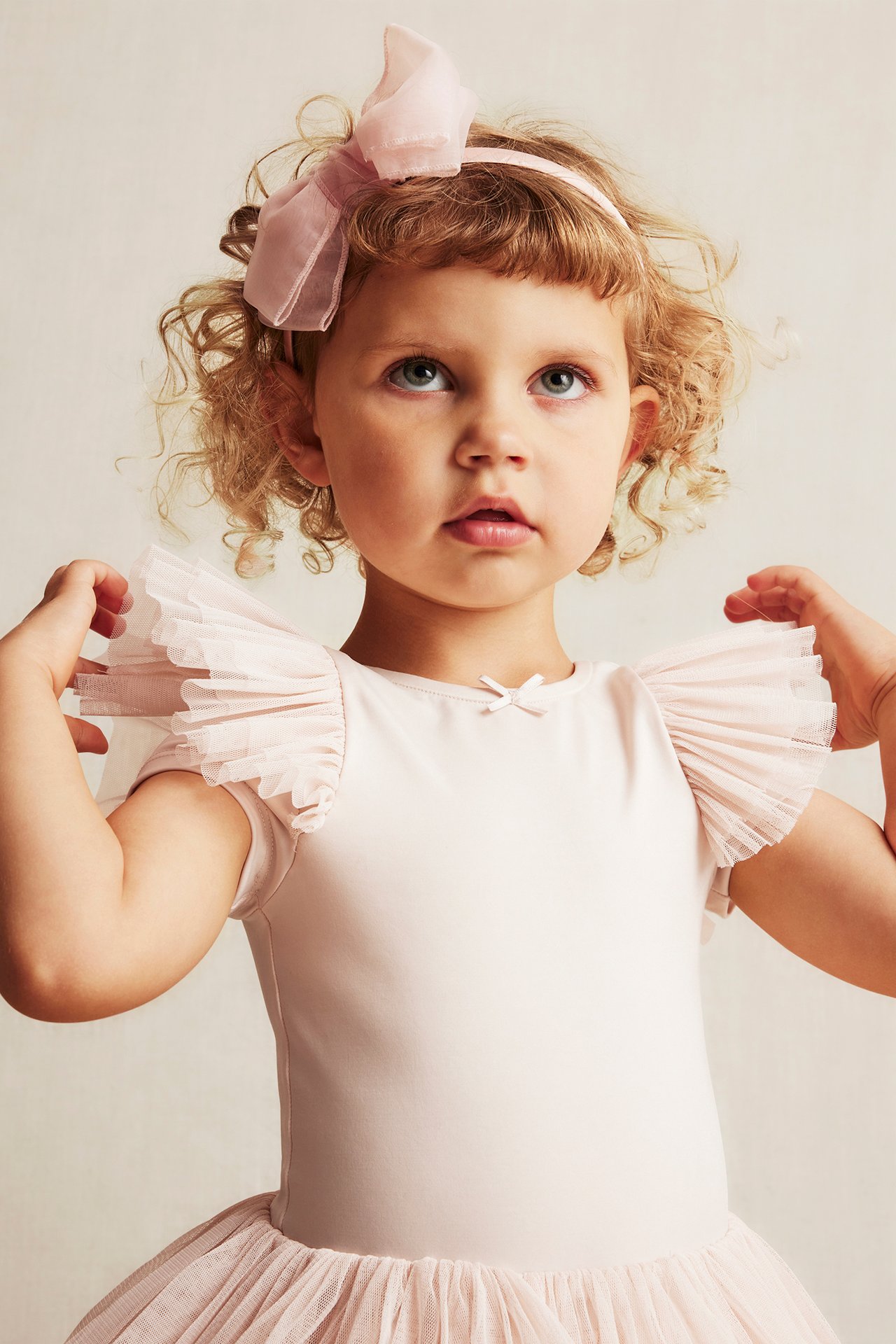 Front view of a kid wearing a pale pink ruffled sleeve ballet leotard, matching tutu, and bow headband.