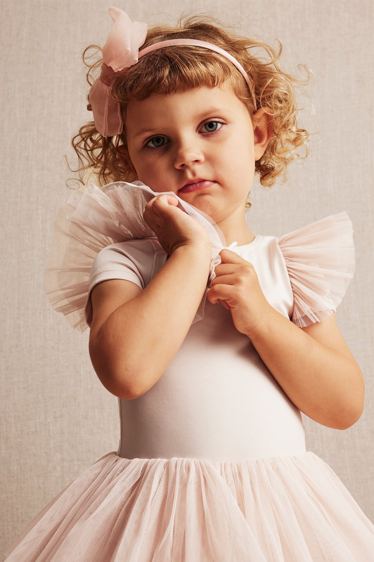 Front view, kid girl in light pink ballerina leotard with mesh ruffles and a pink bow headband.