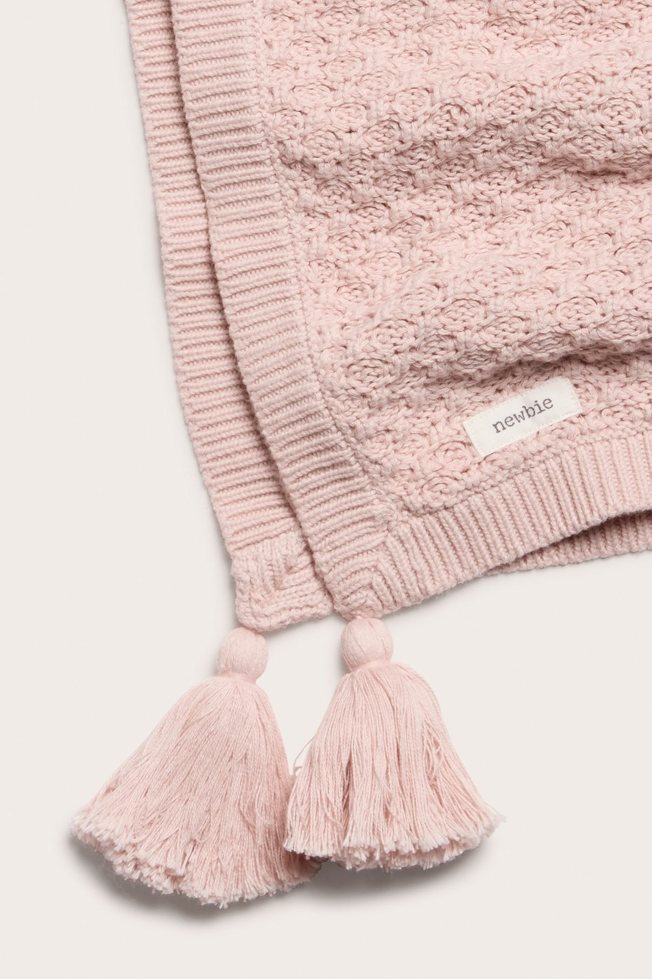 Nahaufnahme einer rosa gemusterten Strick-Babydecke mit Quasten und einem "newbie"-Etikett.
