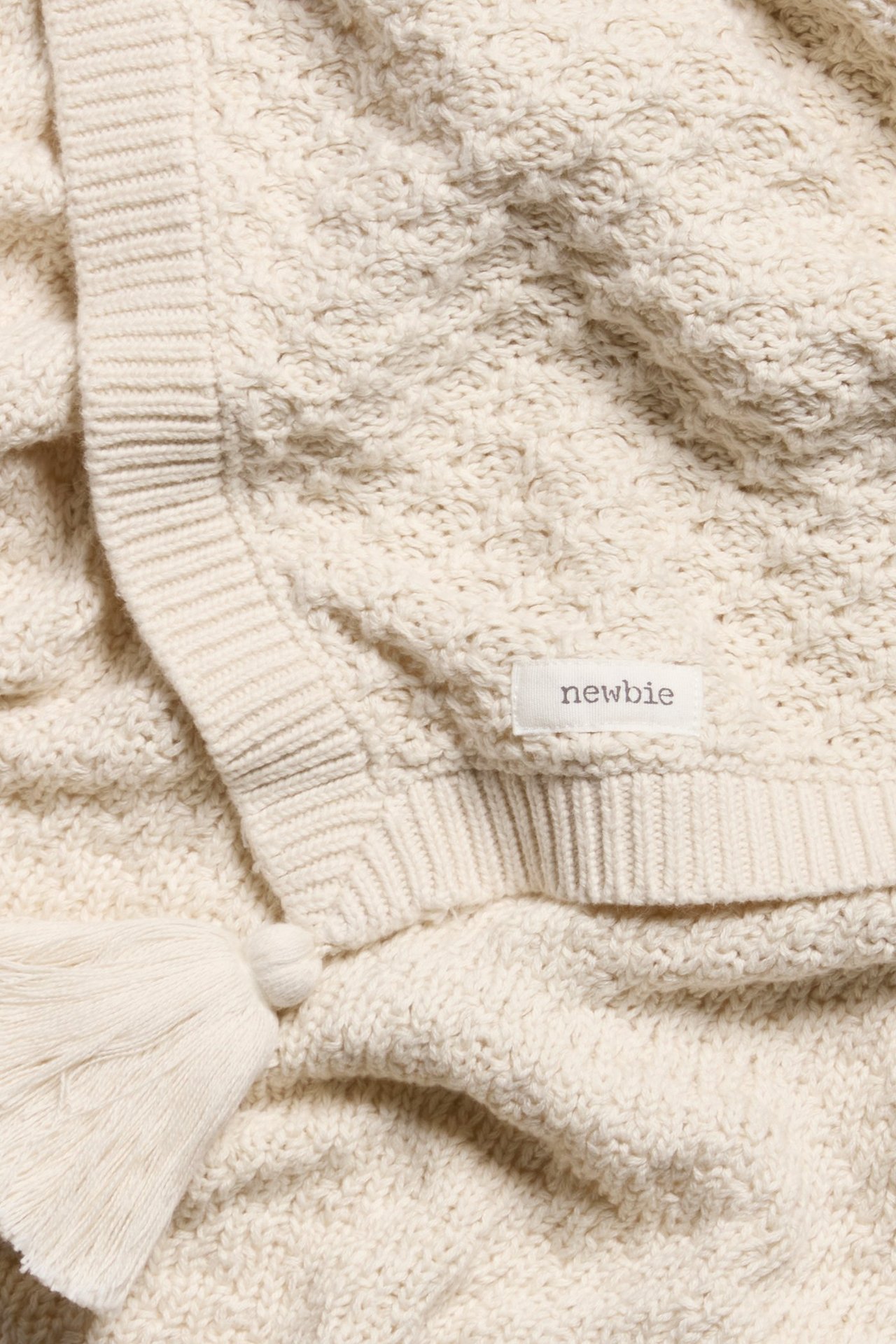 Nahaufnahme einer hellbeigen gemusterten Strick-Babydecke mit Rippenbündchen, Quaste und 'newbie'-Etikett.