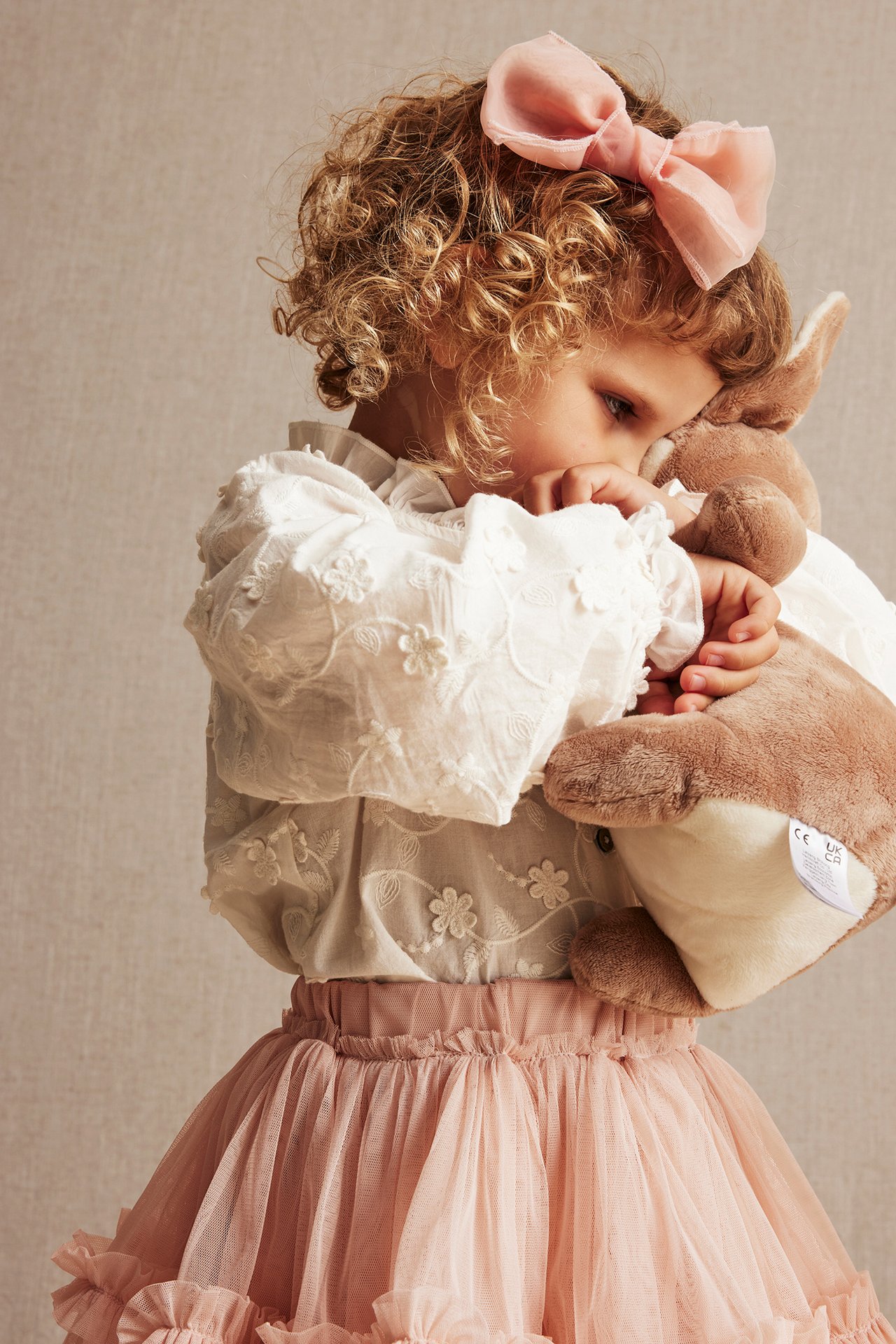 Close-up: Kid in off-white embroidered blouse, pink tulle skirt, pink bow, holding a brown stuffed animal.