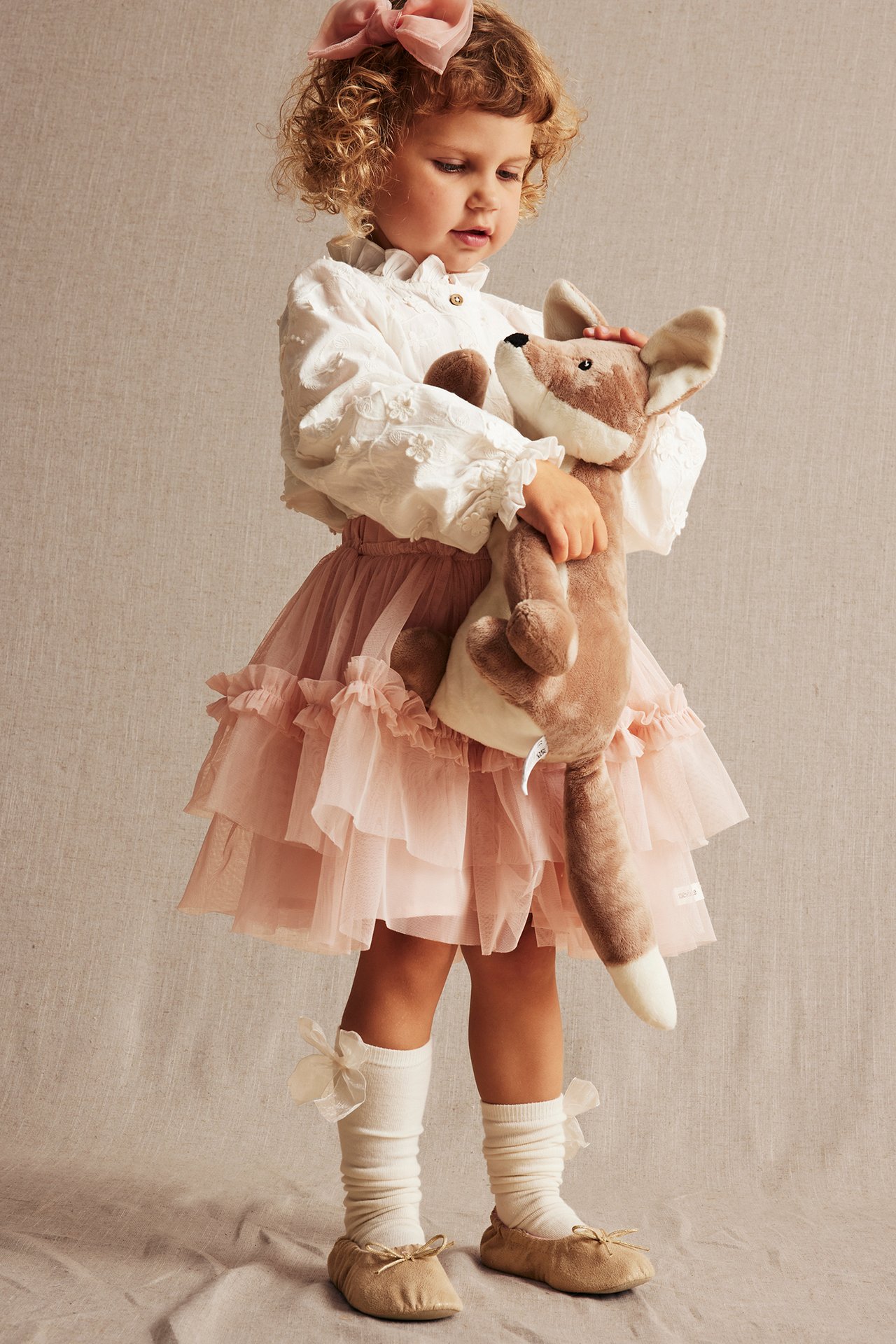 Kid wears off-white embroidered blouse, pink tulle skirt, off-white socks, beige flats, pink bow, holding fox.