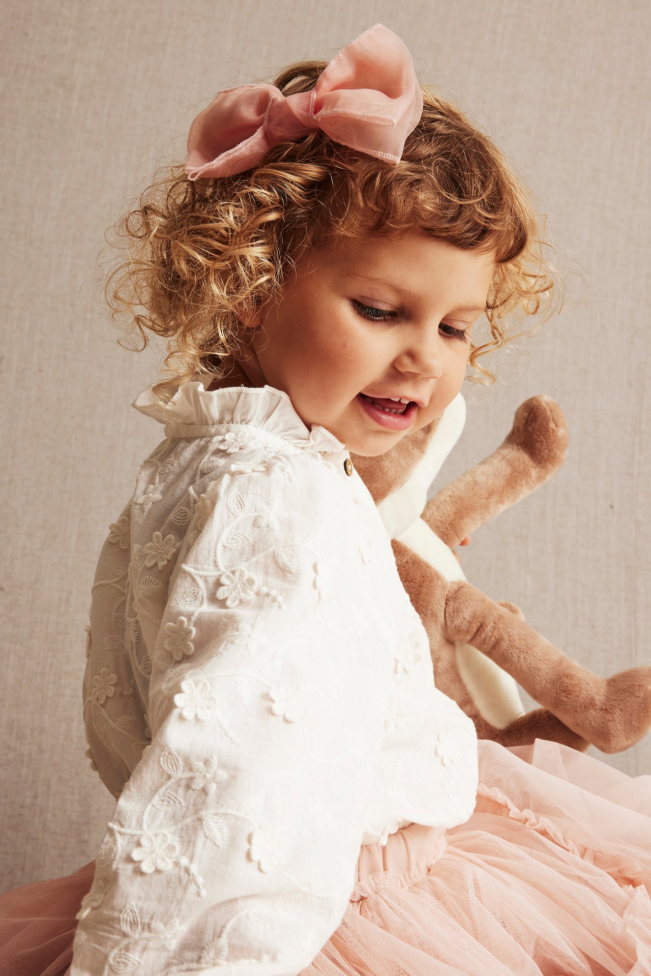 Close-up of a kid in an offwhite embroidered cotton blouse, pink tulle skirt, pink bow, holding a stuffed animal.